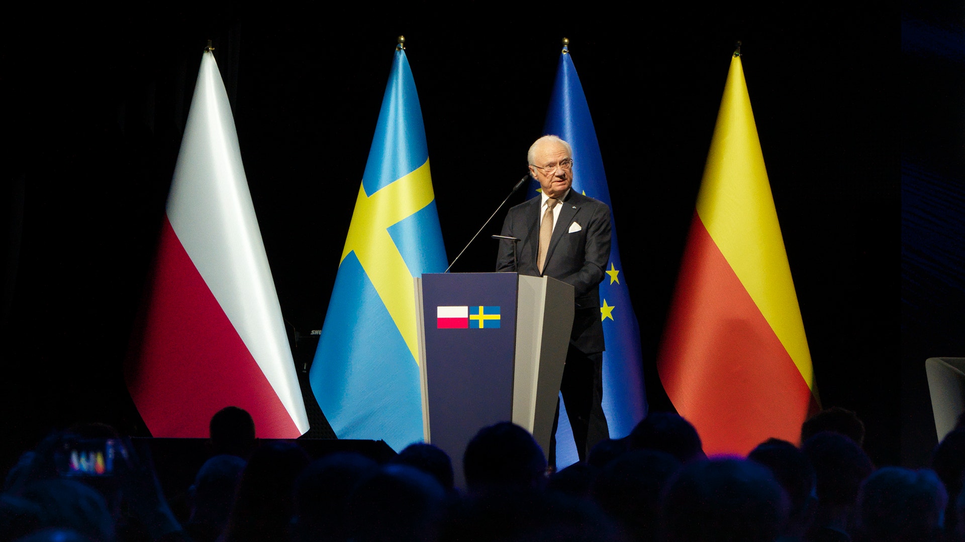 En äldre man talar vid ett podium framför flera flaggor vid en formell ceremoni.