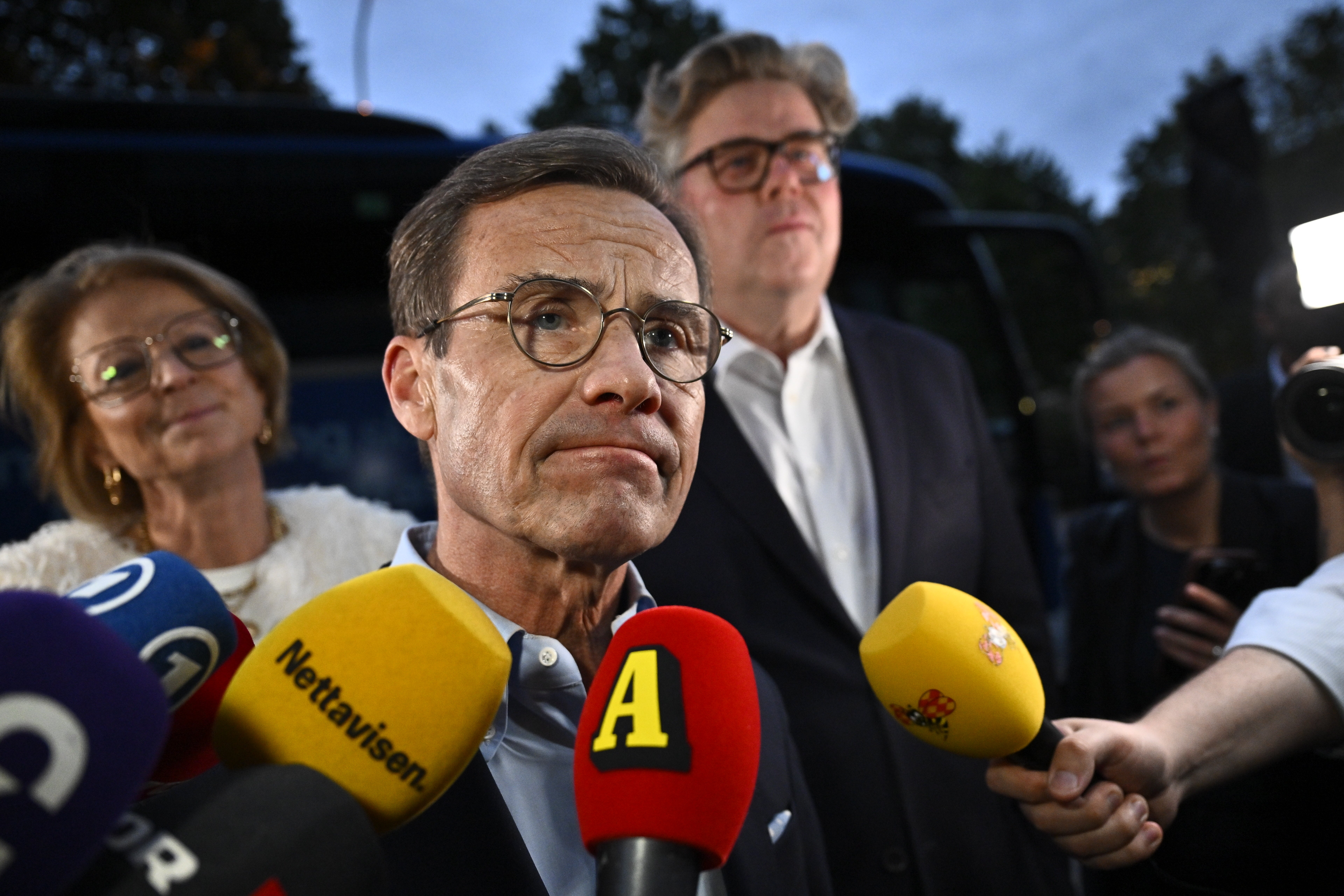 En man i glasögon och kostym intervjuas utomhus av flera journalister som håller fram mikrofoner, med andra personer i bakgrunden.