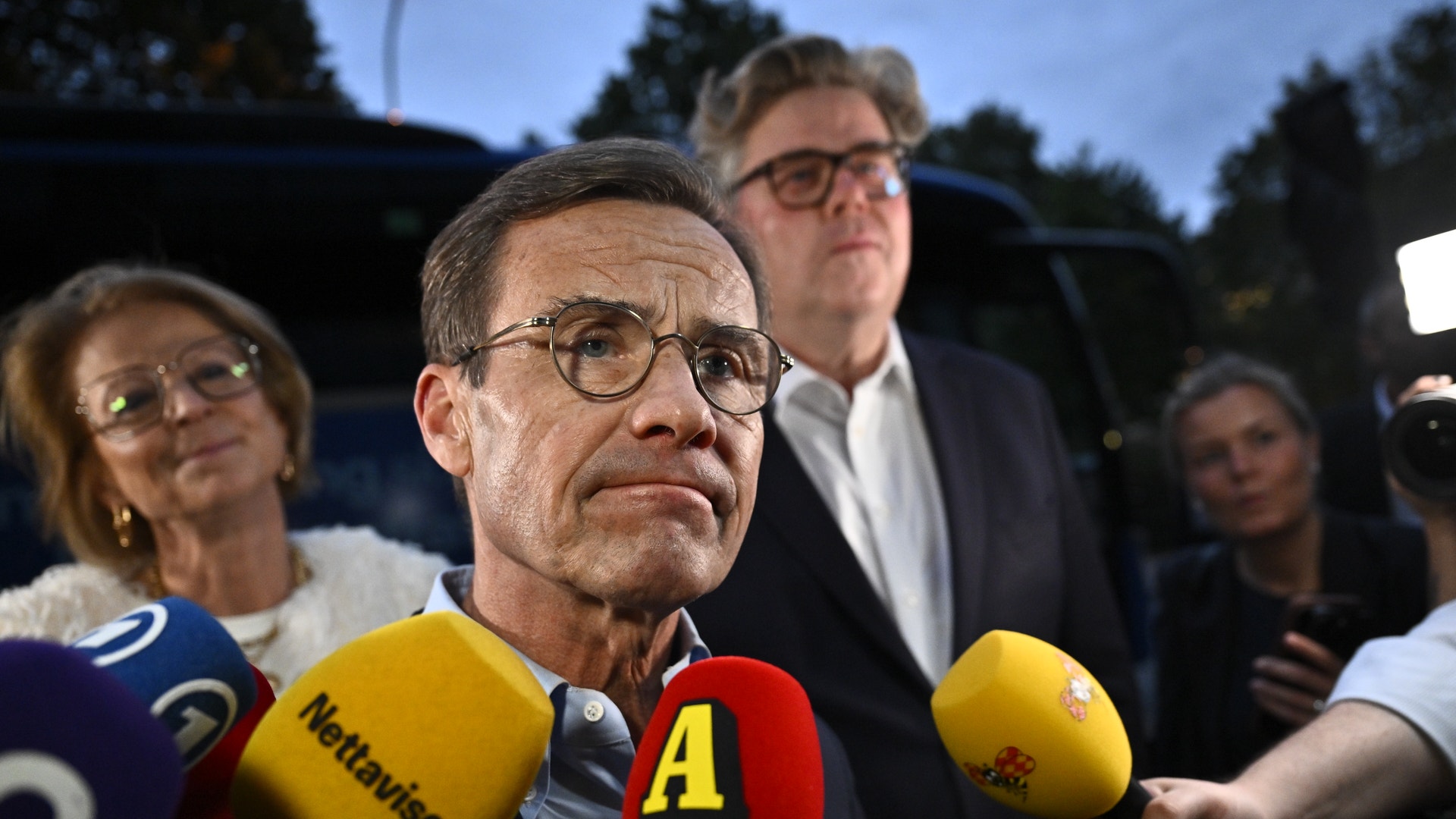 En man i glasögon och kostym intervjuas utomhus av flera journalister som håller fram mikrofoner, med andra personer i bakgrunden.