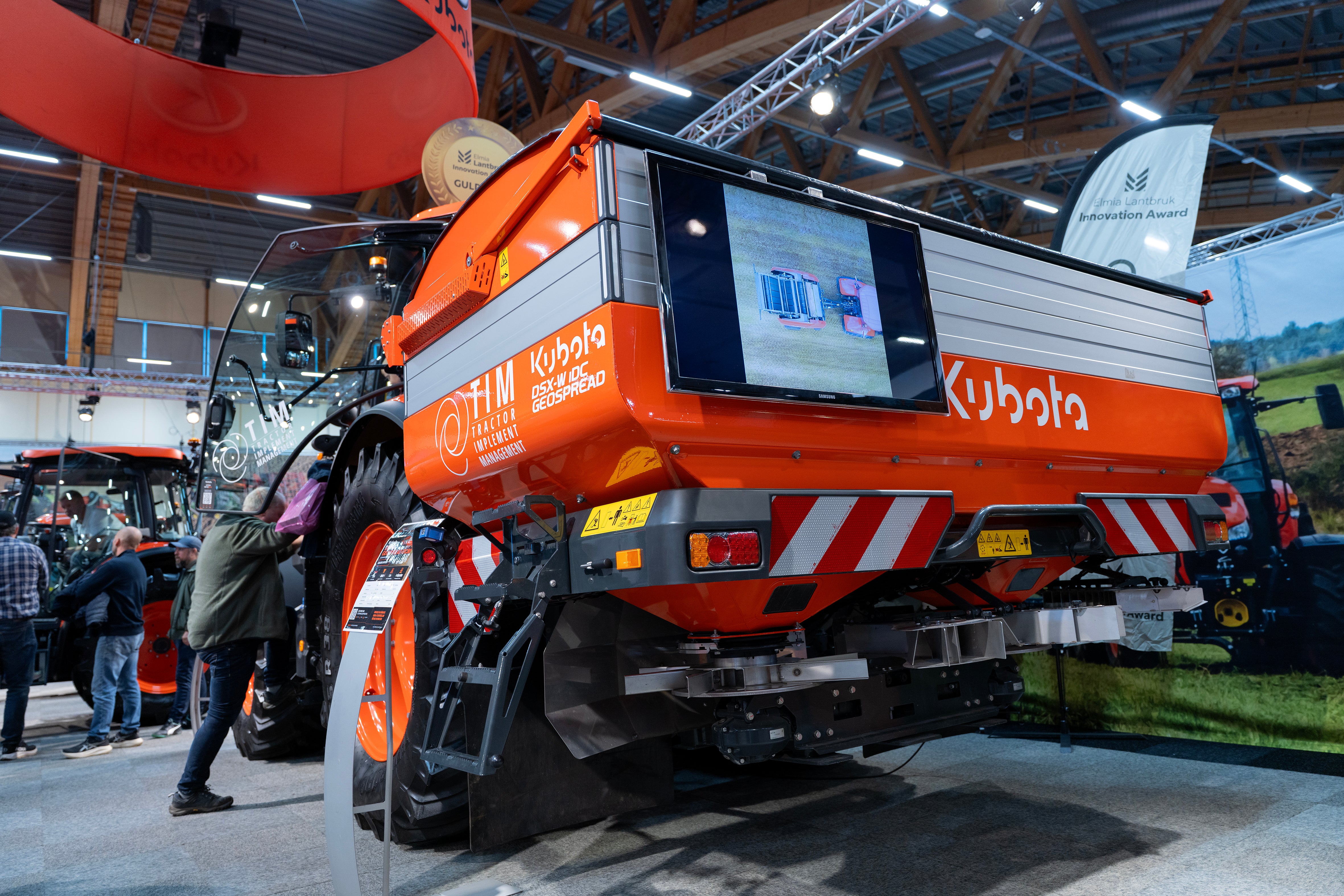 Orange lantbruksmaskin från Kubota på en mässa, med en skärm på baksidan och besökare i bakgrunden.