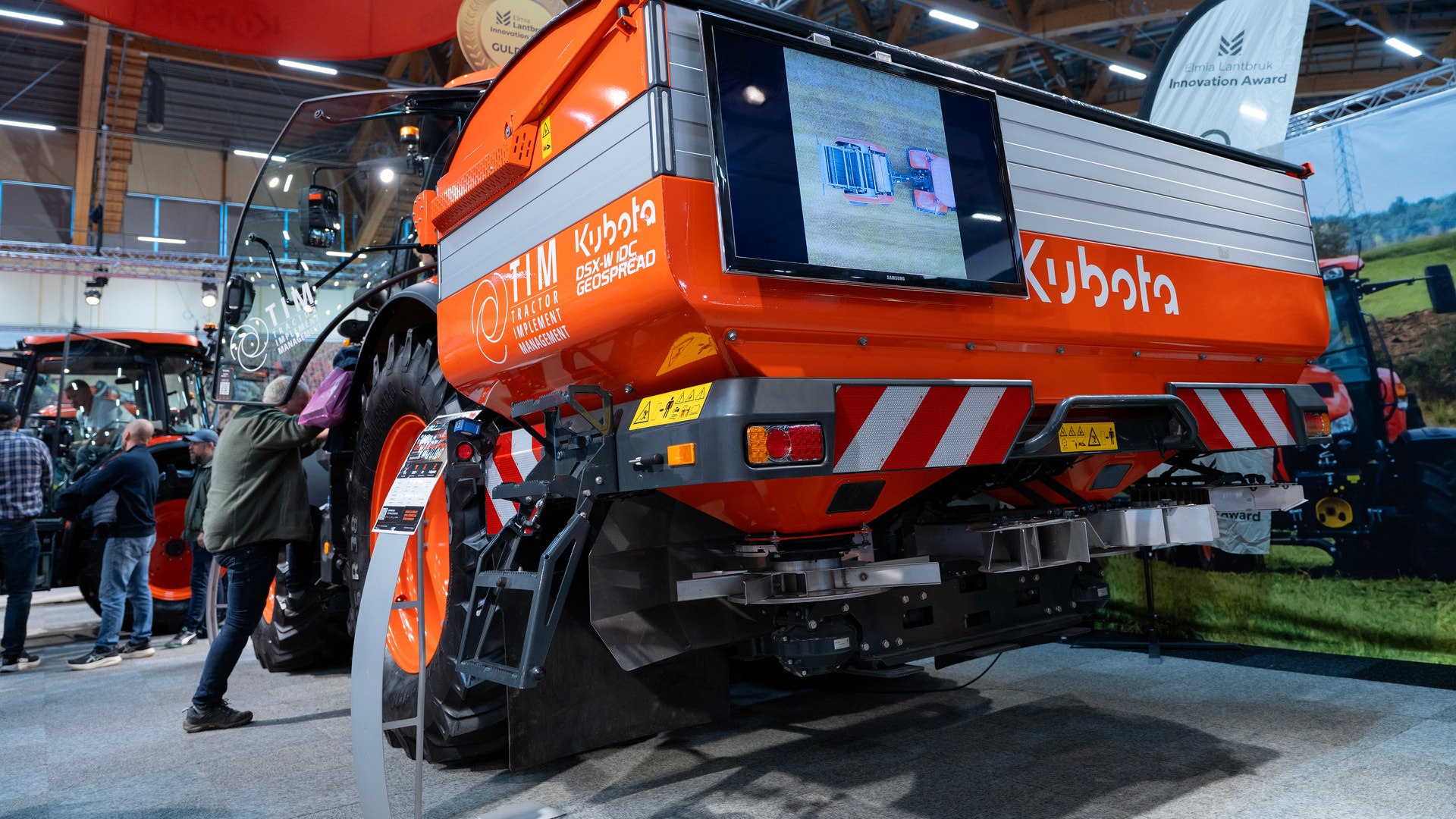 Orange lantbruksmaskin från Kubota på en mässa, med en skärm på baksidan och besökare i bakgrunden.