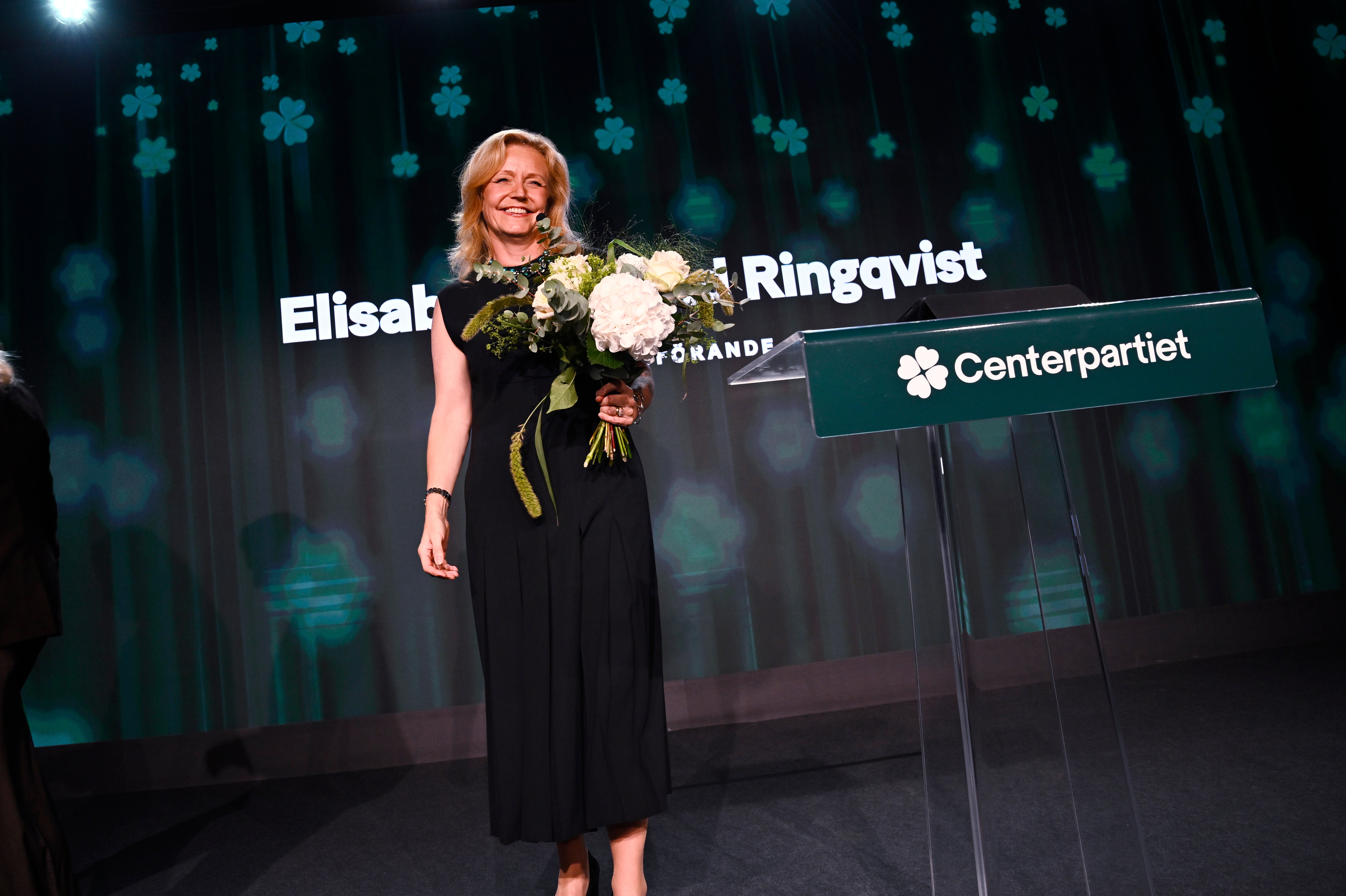 En kvinna i svart klänning står på en scen med en bukett blommor i handen, bredvid en talarstol märkt Centerpartiet.
