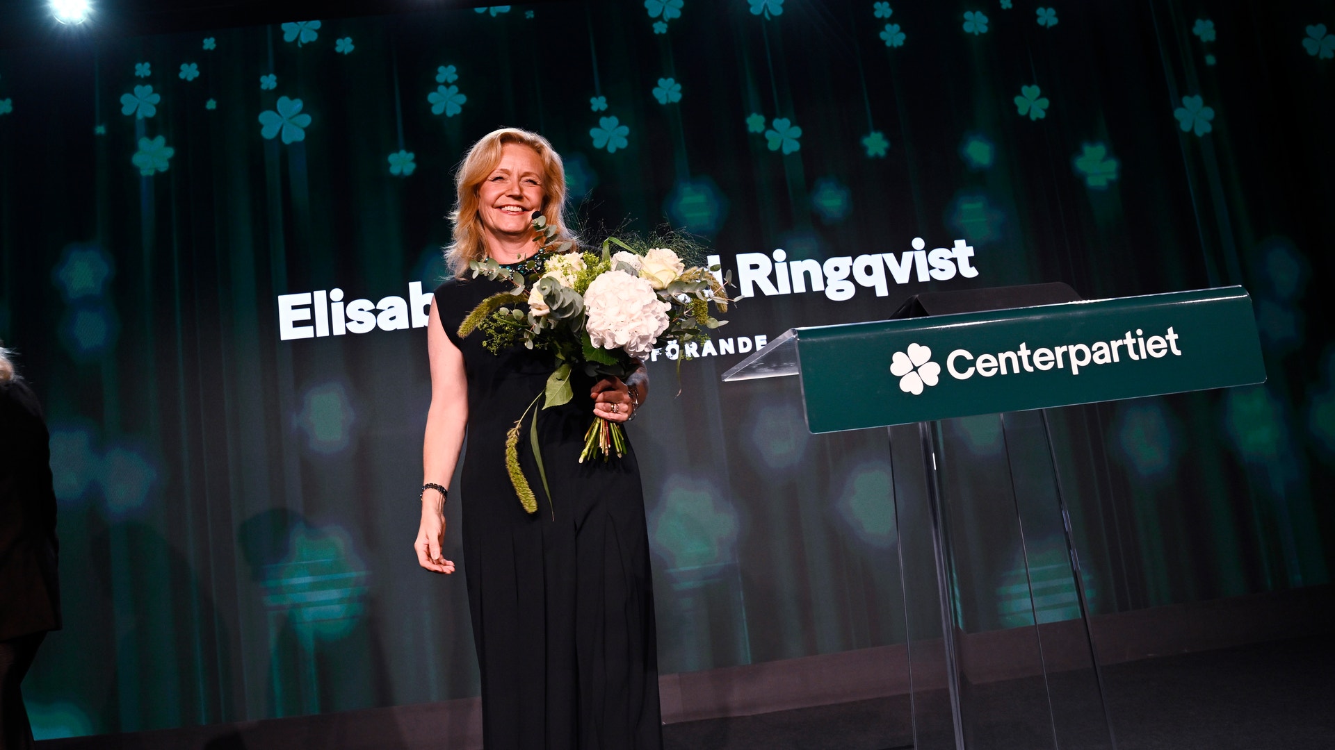 En kvinna i svart klänning står på en scen med en bukett blommor i handen, bredvid en talarstol märkt Centerpartiet.