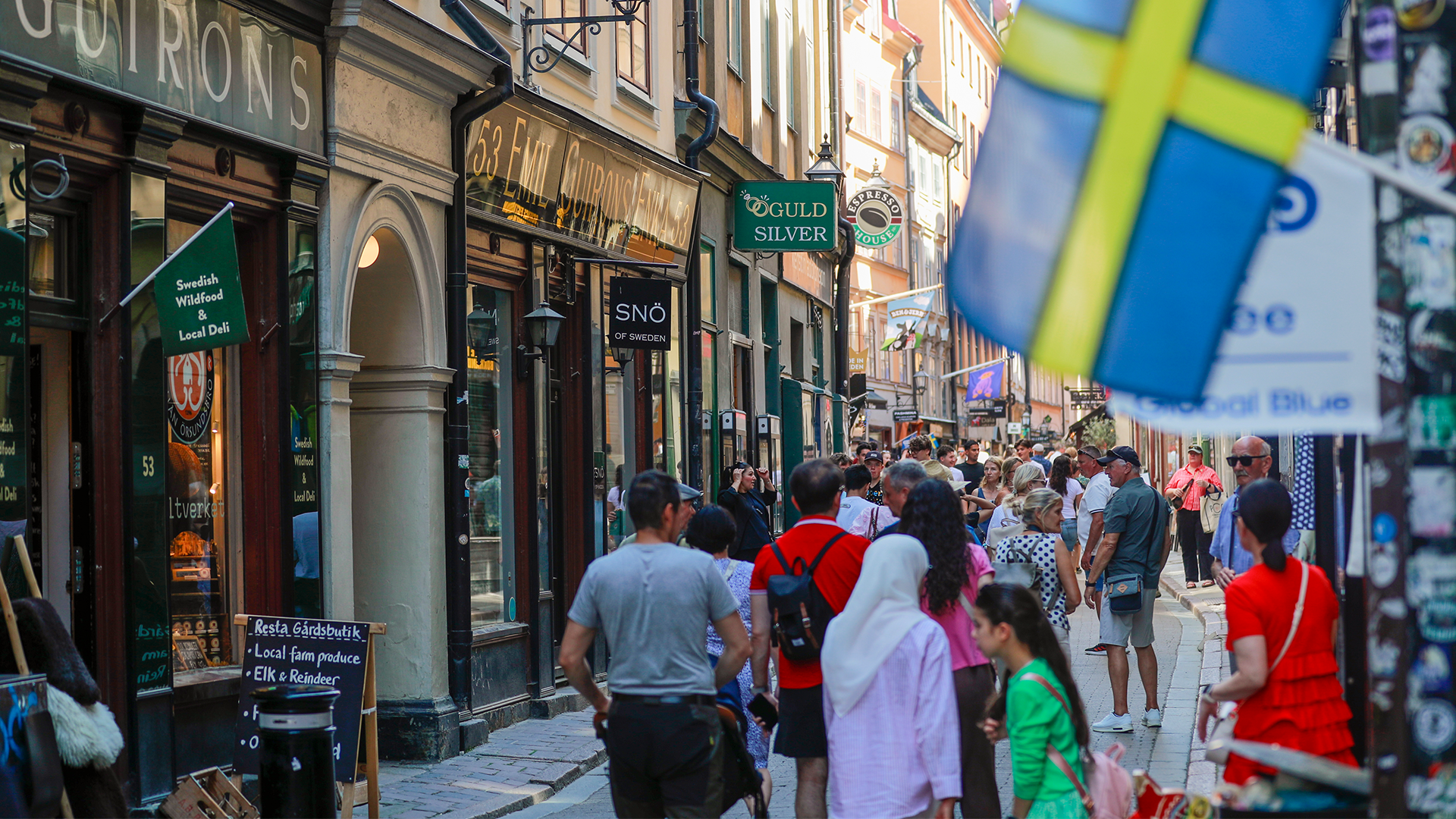 En folkrik gata i Stockholms gamla stad med butiker, restauranger och många turister som promenerar mellan de gamla husfasaderna.