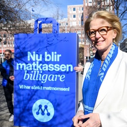 Kvinna i vit blazer och blå scarf håller en blå affisch med texten 'Nu blir matkassen billigare' på en stadsgata.