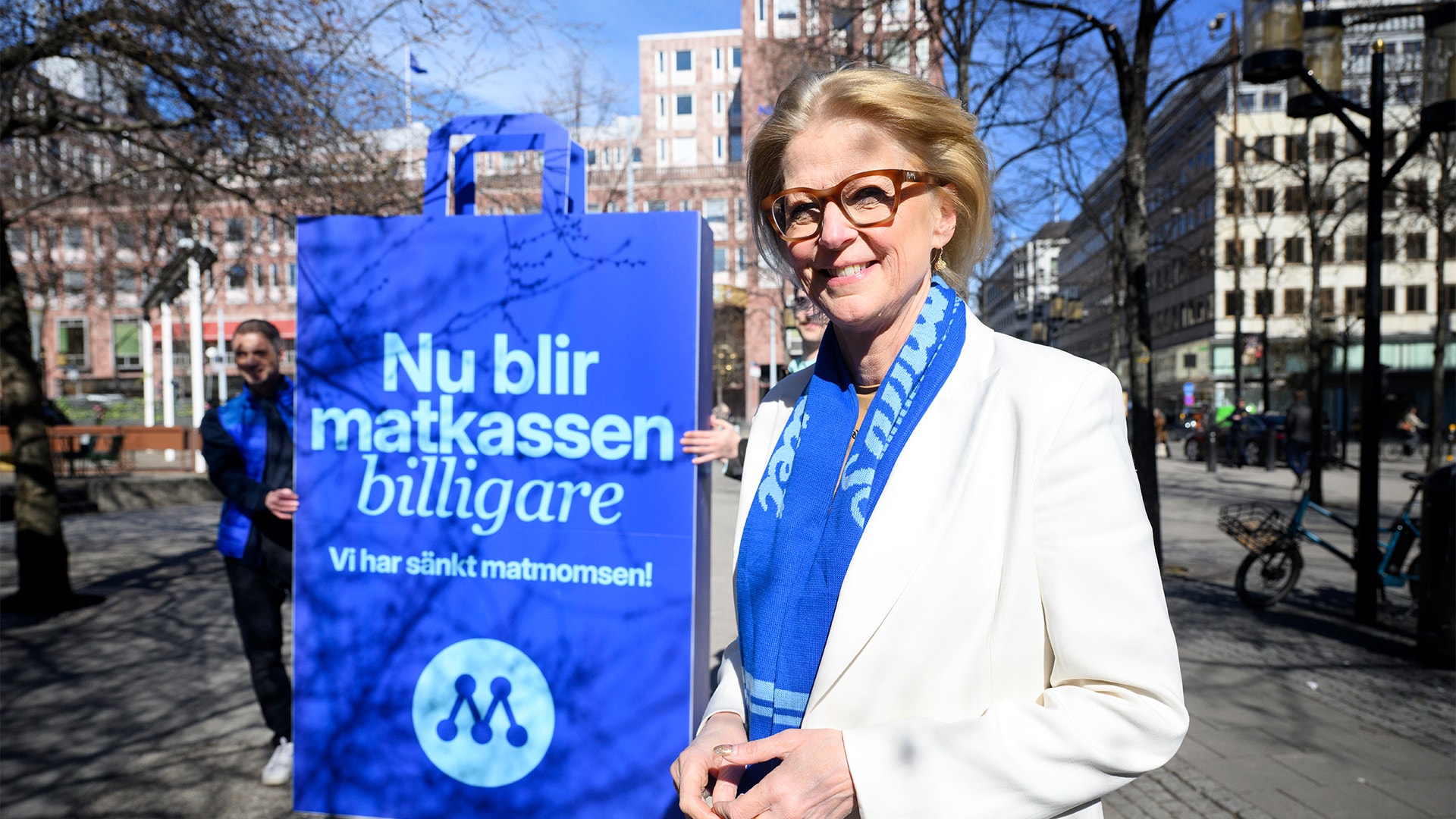 Kvinna i vit blazer och blå scarf håller en blå affisch med texten 'Nu blir matkassen billigare' på en stadsgata.