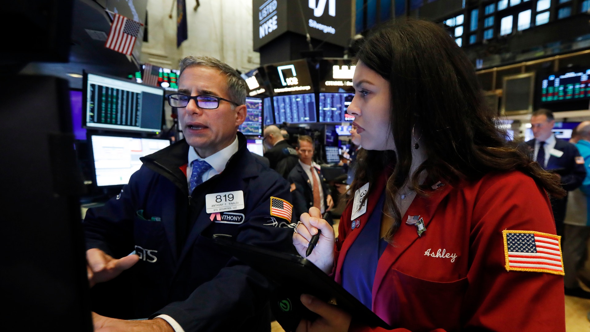 Två handlare på börsgolvet diskuterar framför digitala marknadsvisningar.