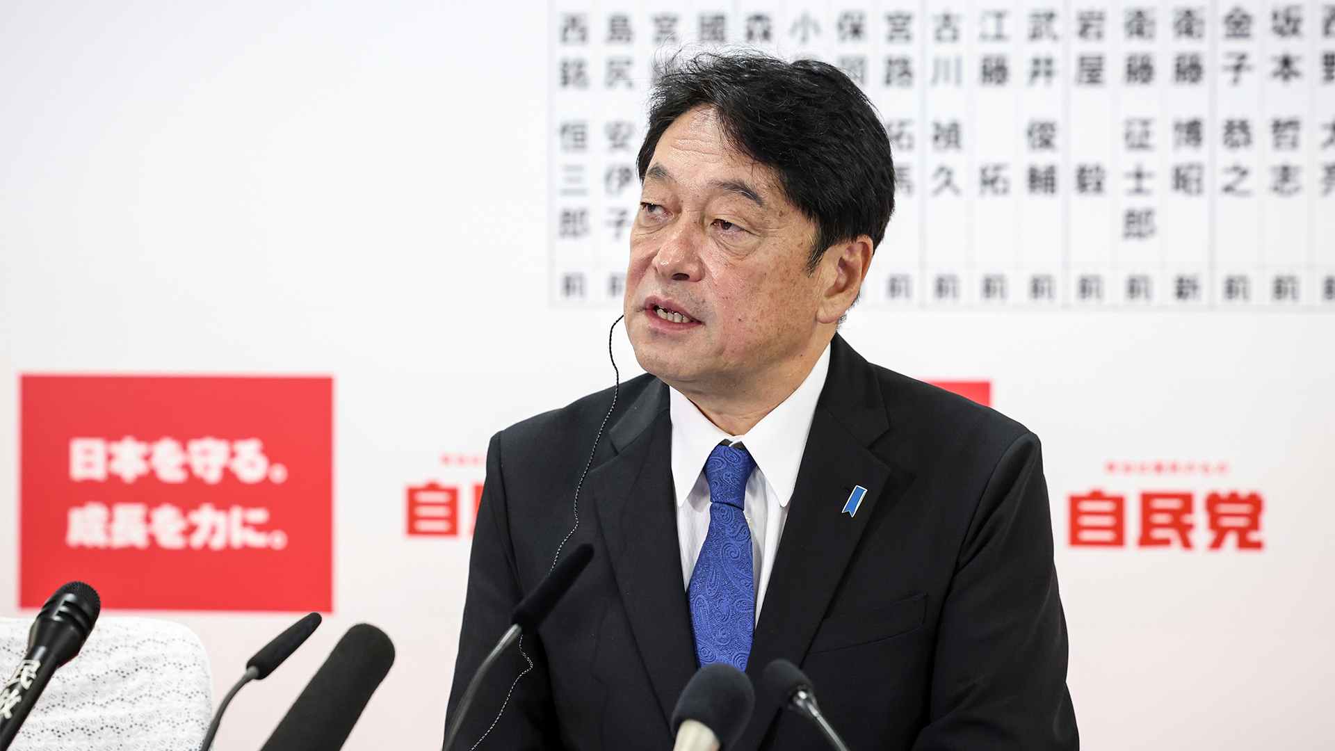 Porträtt av en man i svart kostym och blå slips som talar vid en mikrofon under en presskonferens.