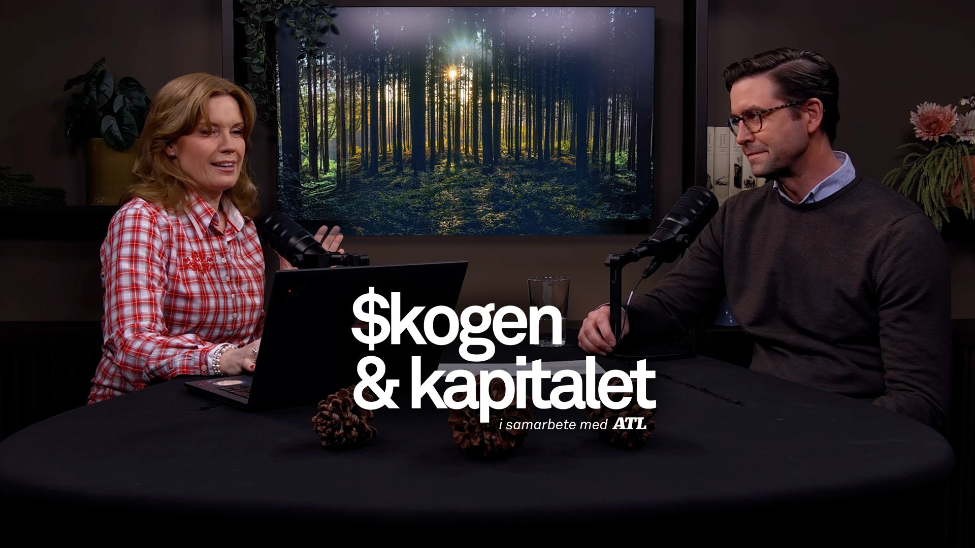 Två personer sitter vid ett bord och samtalar i en studiomiljö med en skogsbild på en skärm bakom dem.