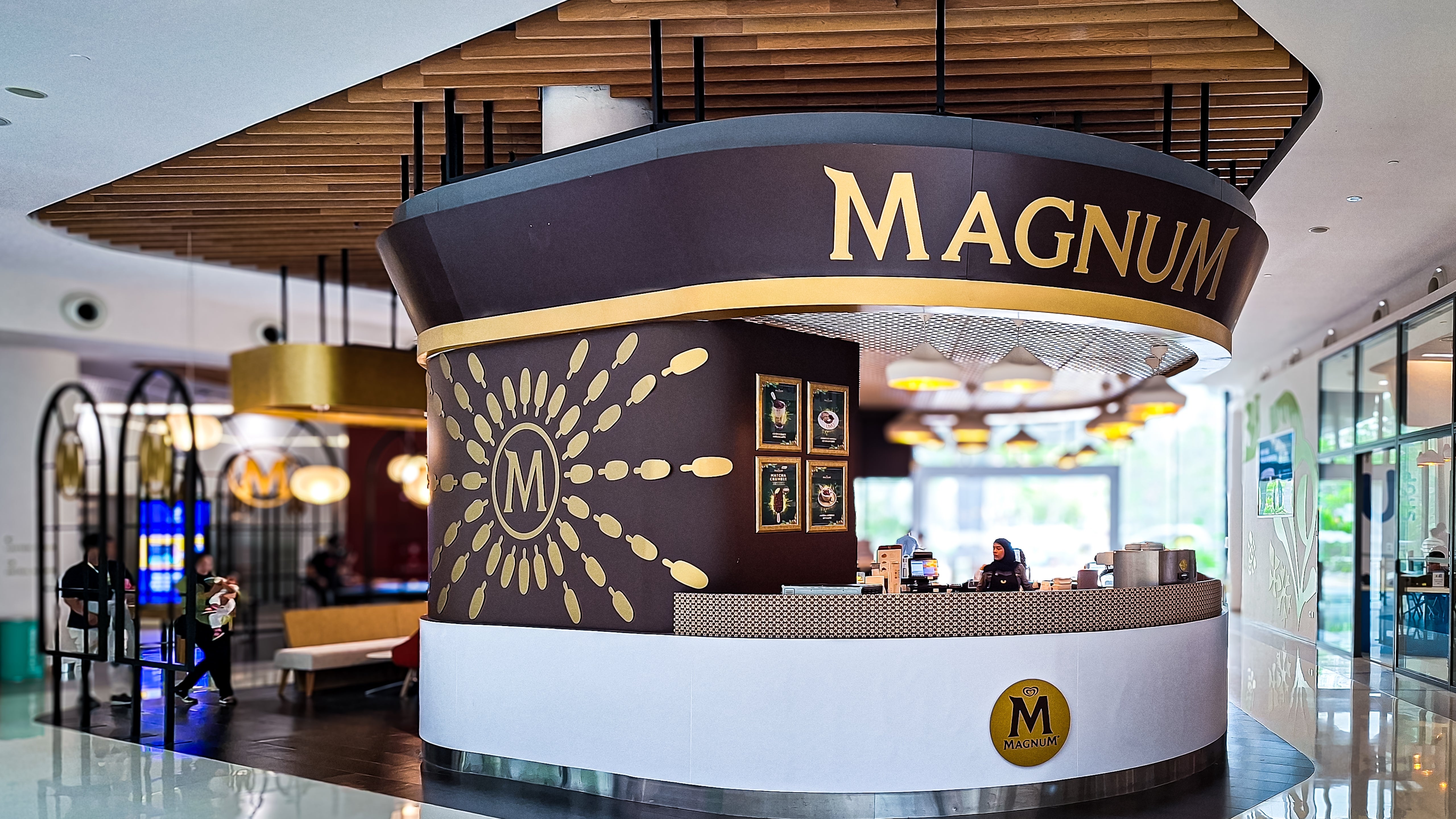 En rundformad Magnum-kiosk i ett köpcenter med svart och vit design, gulddetaljer och en solmotiv-logotyp på framsidan.