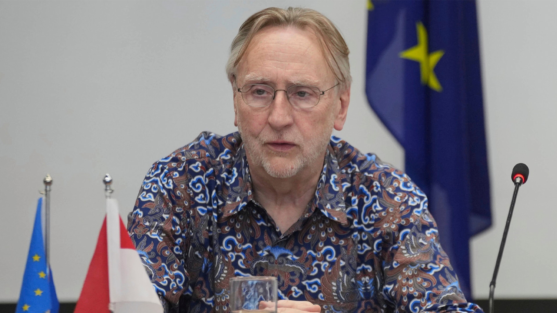 Porträtt av en man med glasögon som talar vid ett officiellt evenemang med flaggor i bakgrunden.