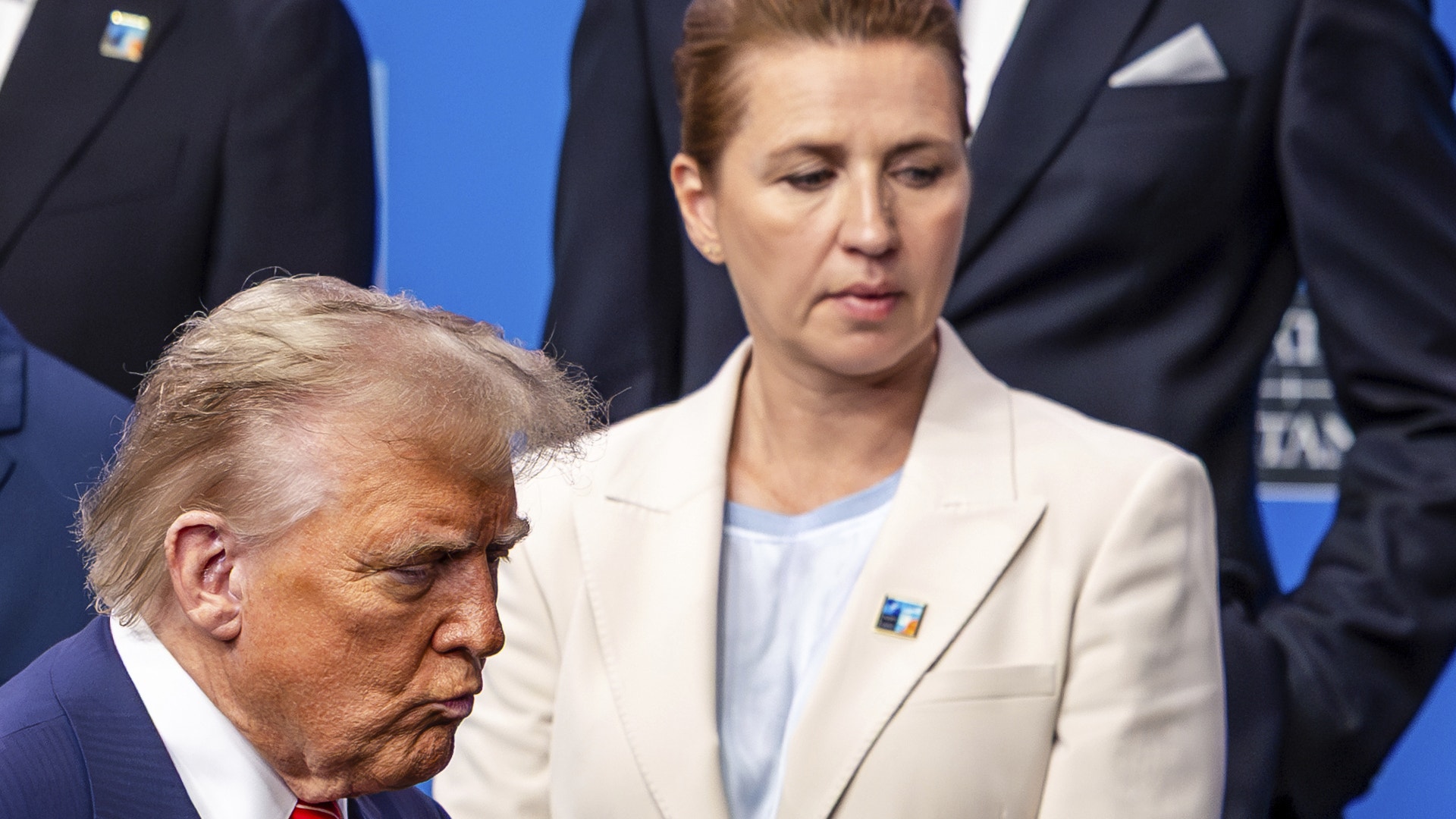 Donald Trump i profil tillsammans med en kvinna i beige kostym vid en officiell händelse framför en blå bakgrund. Trump bär mörk kostym med röd slips. I bakgrunden syns andra människor i formell klädsel.
