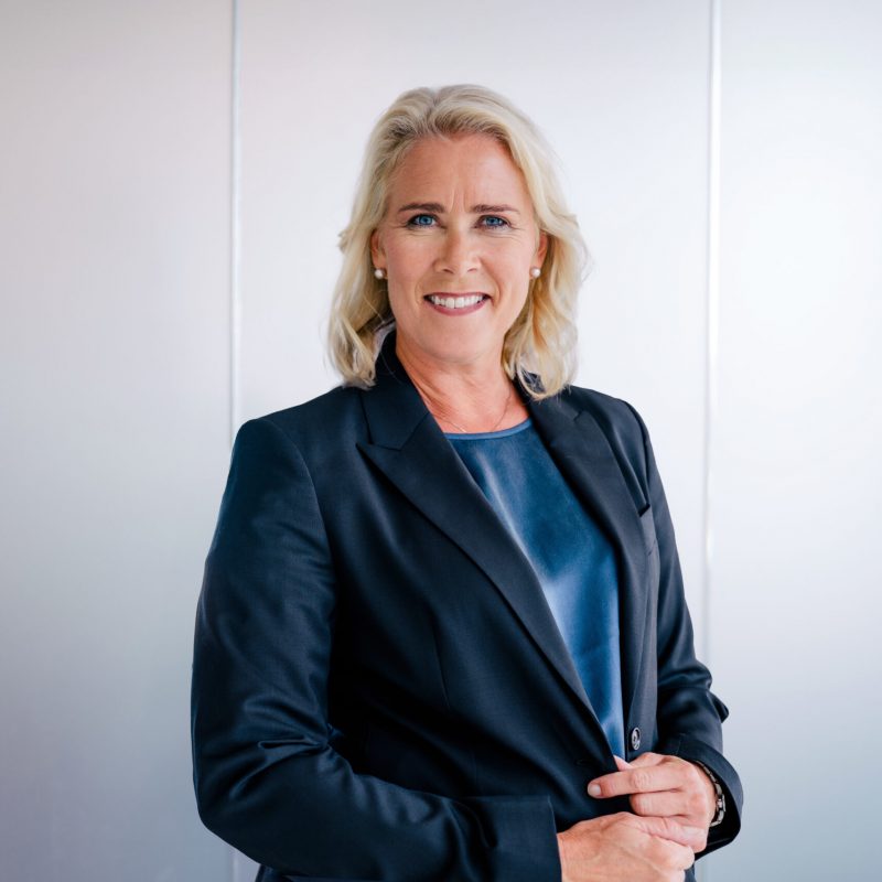 Porträtt av en kvinna med blont hår som ler mot kameran. Hon bär en mörkblå kavaj och blå skjorta framför en vit vägg.
