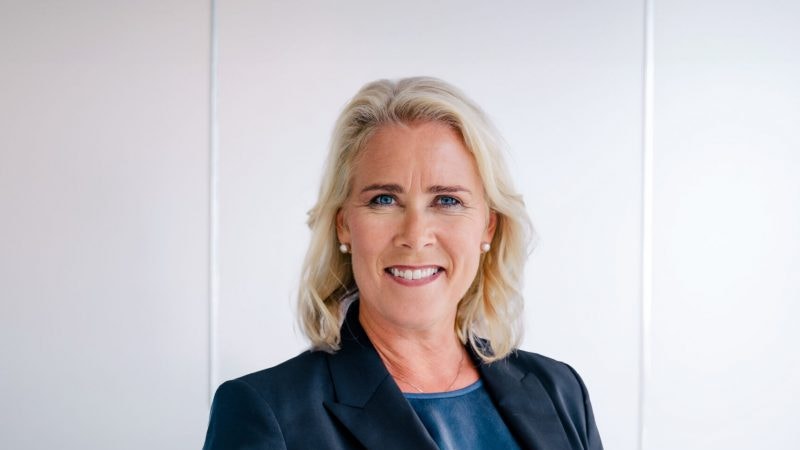 Porträtt av en kvinna med blont hår som ler mot kameran. Hon bär en mörkblå kavaj och blå skjorta framför en vit vägg.