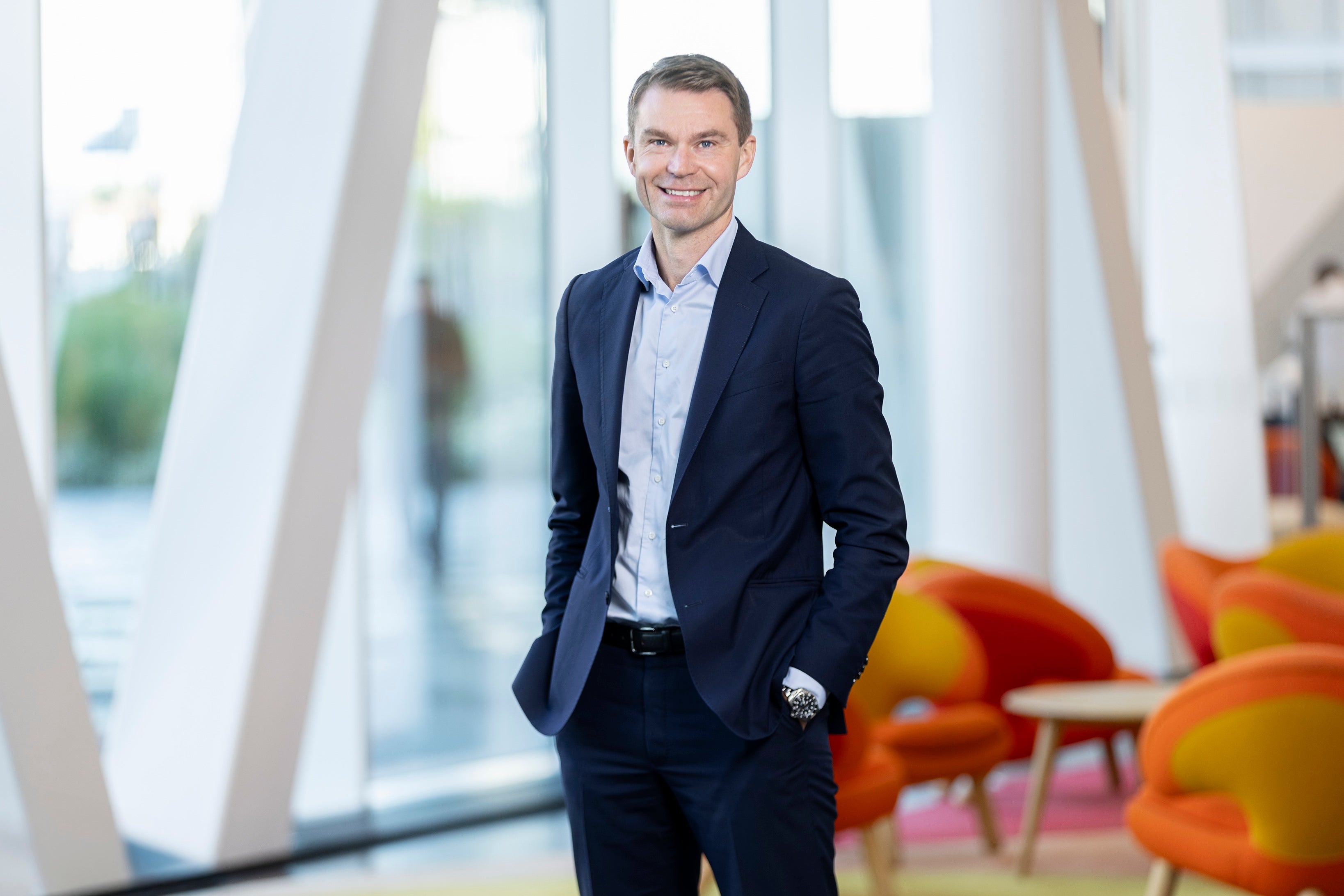 Porträtt av en man i mörkblå kostym och ljusblå skjorta som står i en modern kontorsmiljö med stora fönster.