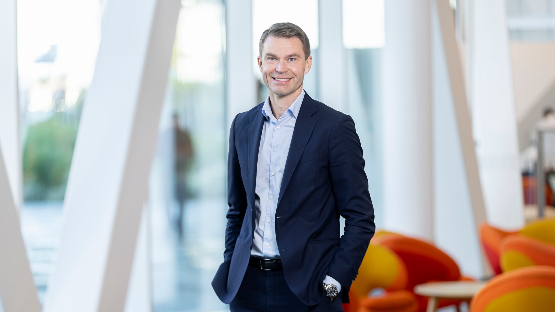 Porträtt av en man i mörkblå kostym och ljusblå skjorta som står i en modern kontorsmiljö med stora fönster.
