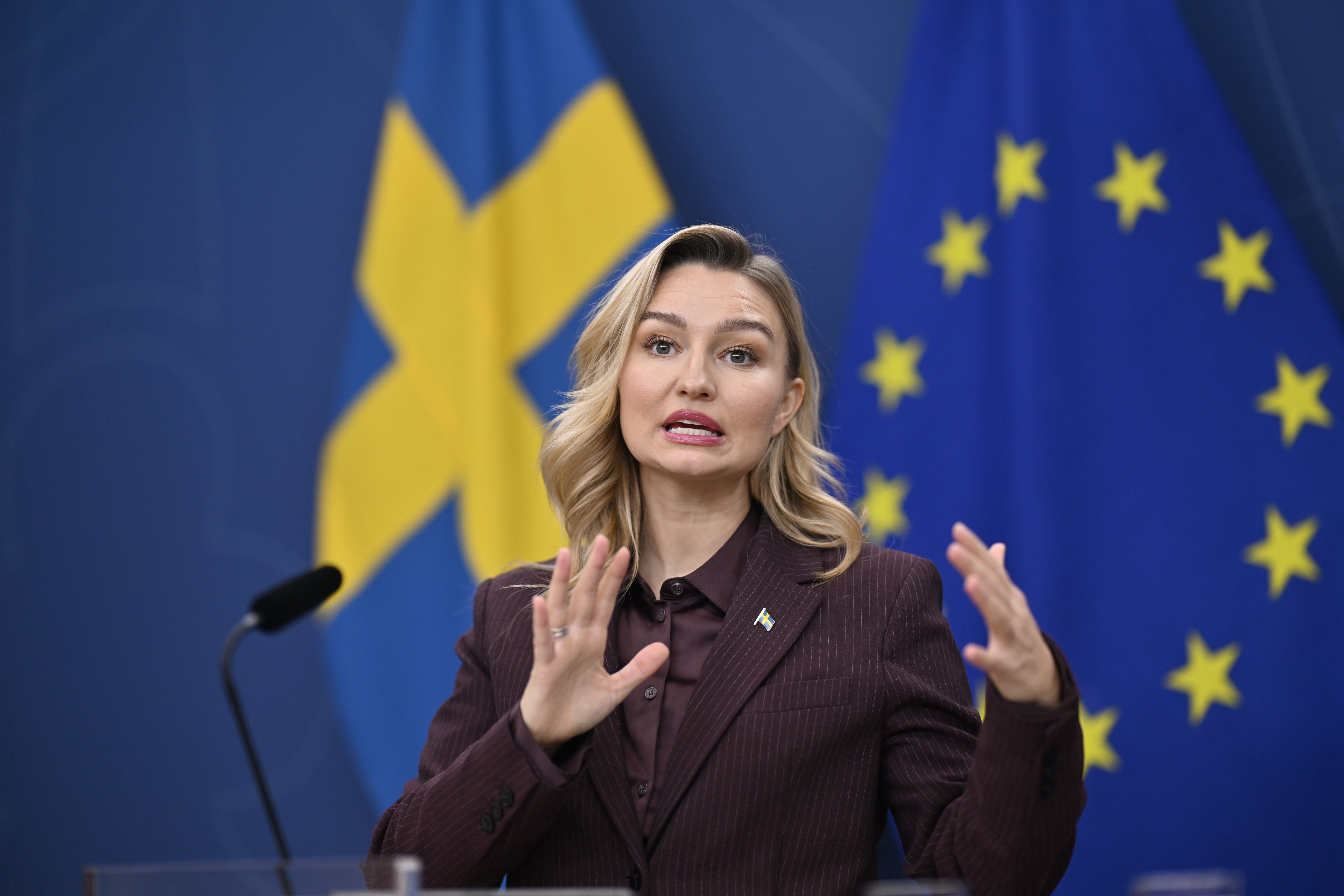 Kvinna i brun kostym gestikulerar med båda händerna när hon talar vid en presskonferens, framför en svensk flagga och en EU-flagga.