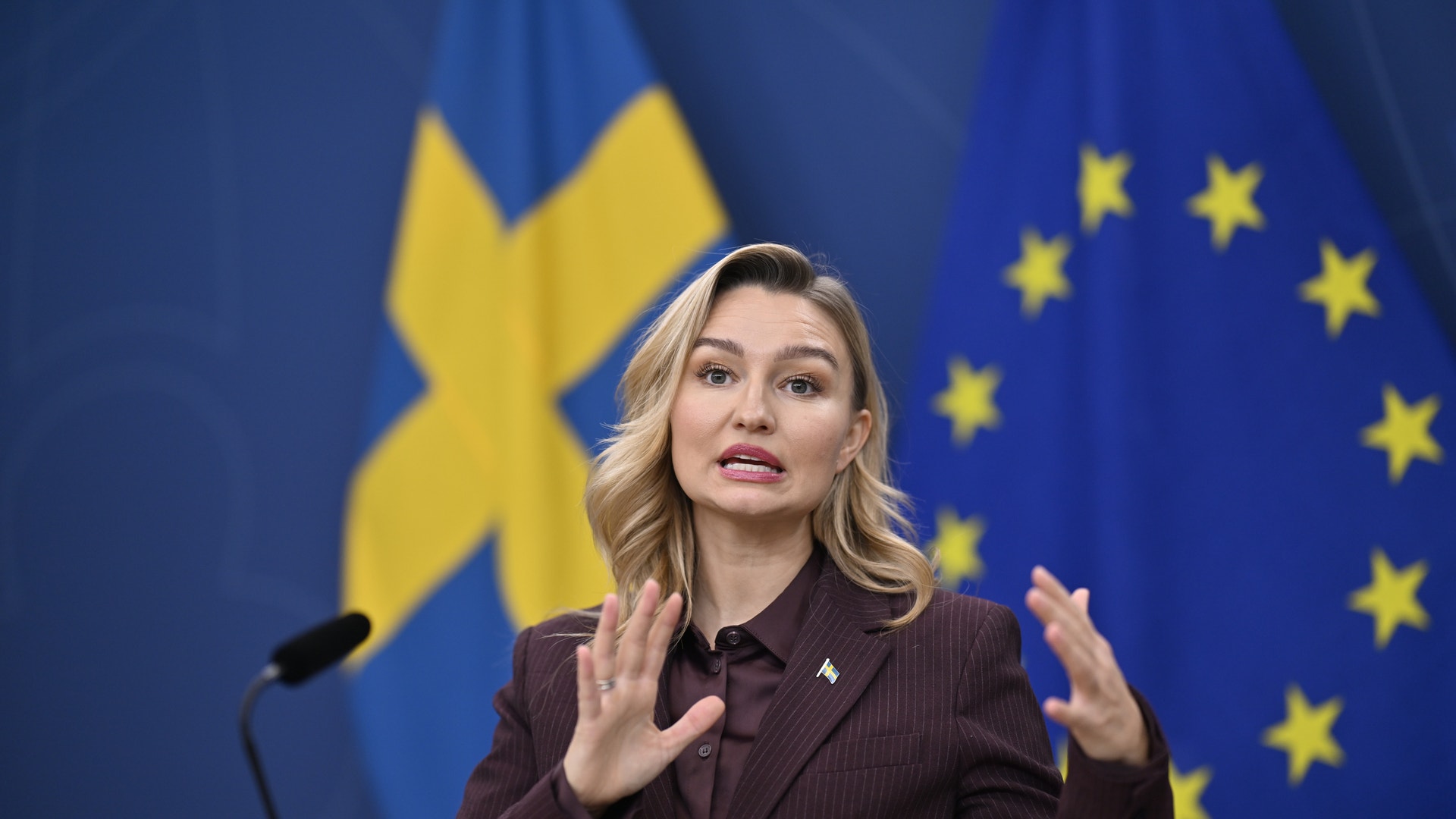 Kvinna i brun kostym gestikulerar med båda händerna när hon talar vid en presskonferens, framför en svensk flagga och en EU-flagga.