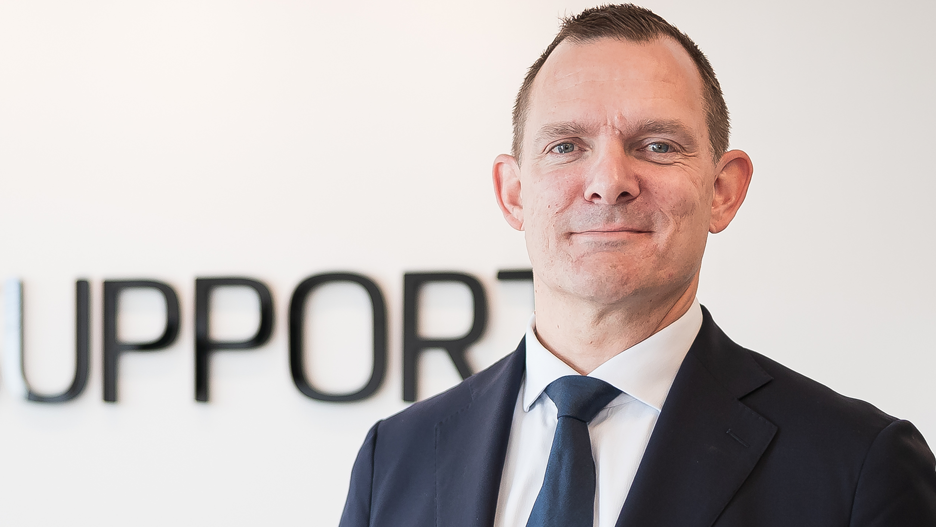 Porträtt av en man i mörkblå kostym och blå slips mot en ljus bakgrund.