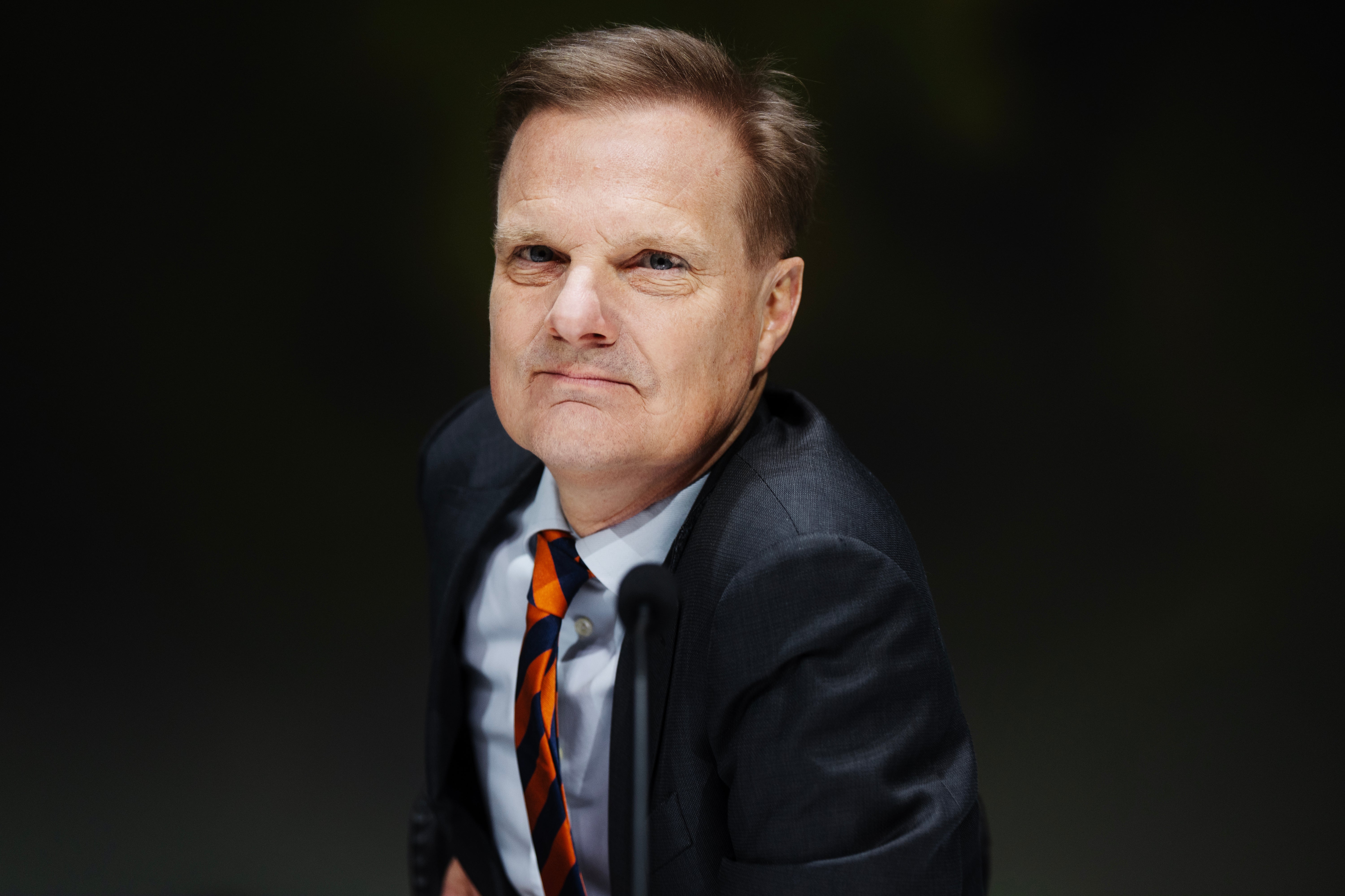 Porträttfoto av en man i mörkblå kostym och orange slips mot mörk bakgrund.