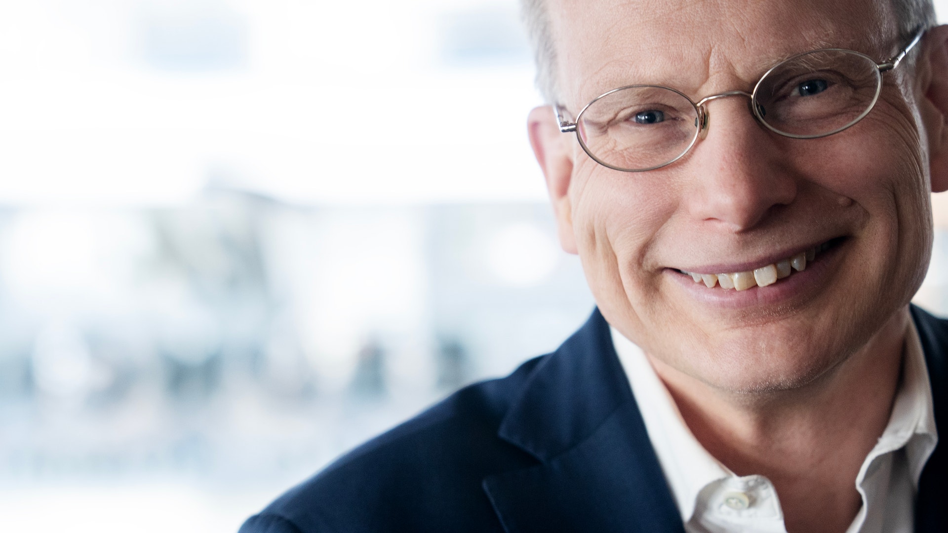 Porträtt av en man med glasögon som ler mot kameran. Han bär en mörkblå kavaj och vit skjorta.