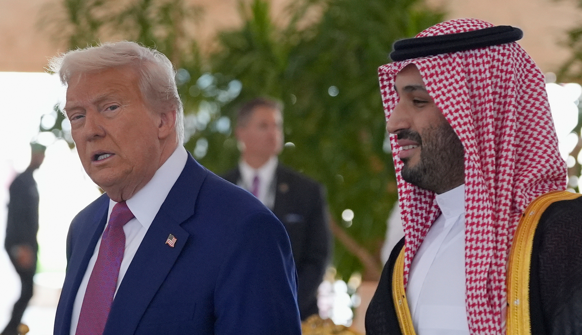 En man i kostym och slips går bredvid en man klädd i traditionell arabisk dräkt med rutig huvudduk, utomhus i solljus.
