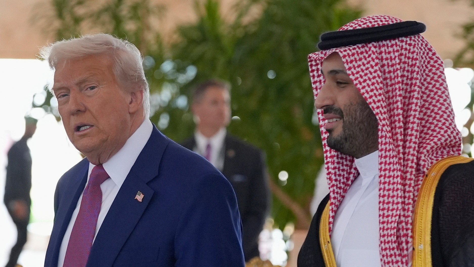 En man i kostym och slips går bredvid en man klädd i traditionell arabisk dräkt med rutig huvudduk, utomhus i solljus.