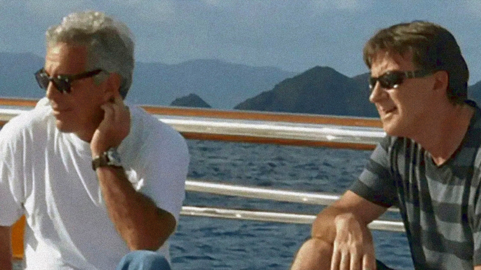 Jeffrey Esptein och Peter Mandelsonsitter på däck på en båt och samtalar. Epstein till vänster bär vita solglasögon och en vit t-shirt, Mandelson till höger bär mörka solglasögon och en grå t-shirt. I bakgrunden syns ett vackert kustlandskap med blått hav och bergiga öar under klar himmel.