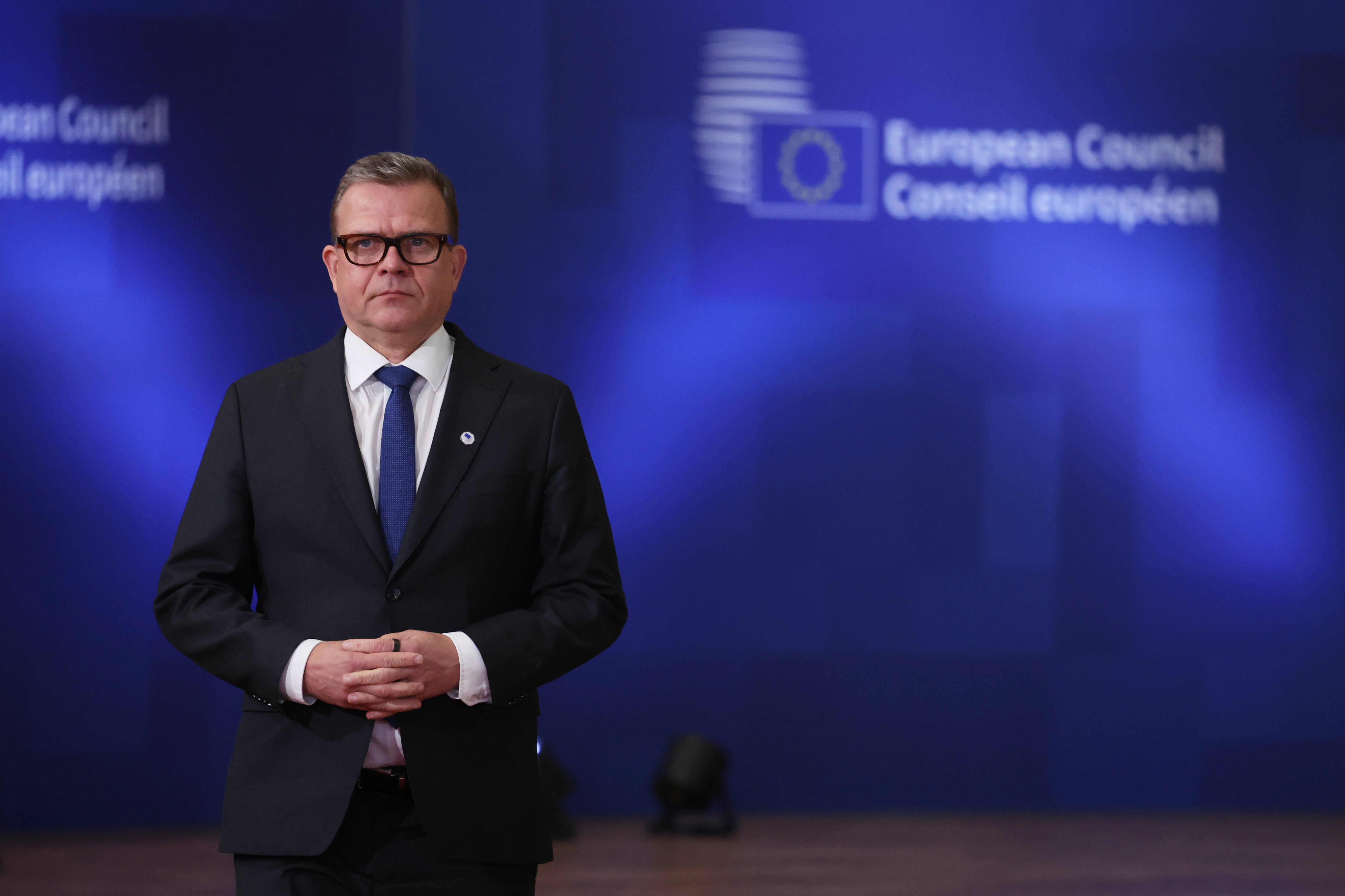 En man i svart kostym och glasögon står framför en blå bakgrund med Europarådet logotyp.