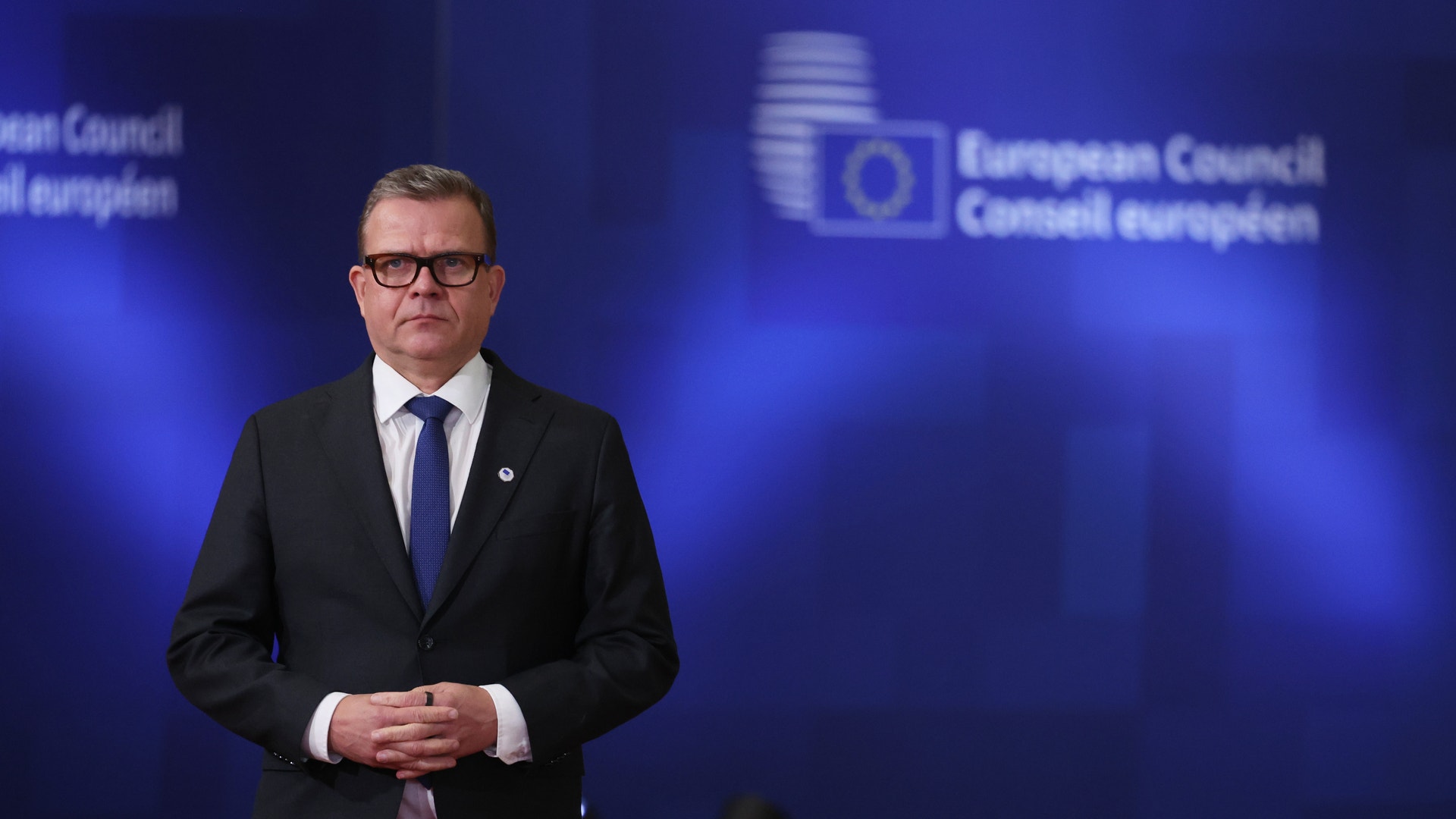 En man i svart kostym och glasögon står framför en blå bakgrund med Europarådet logotyp.