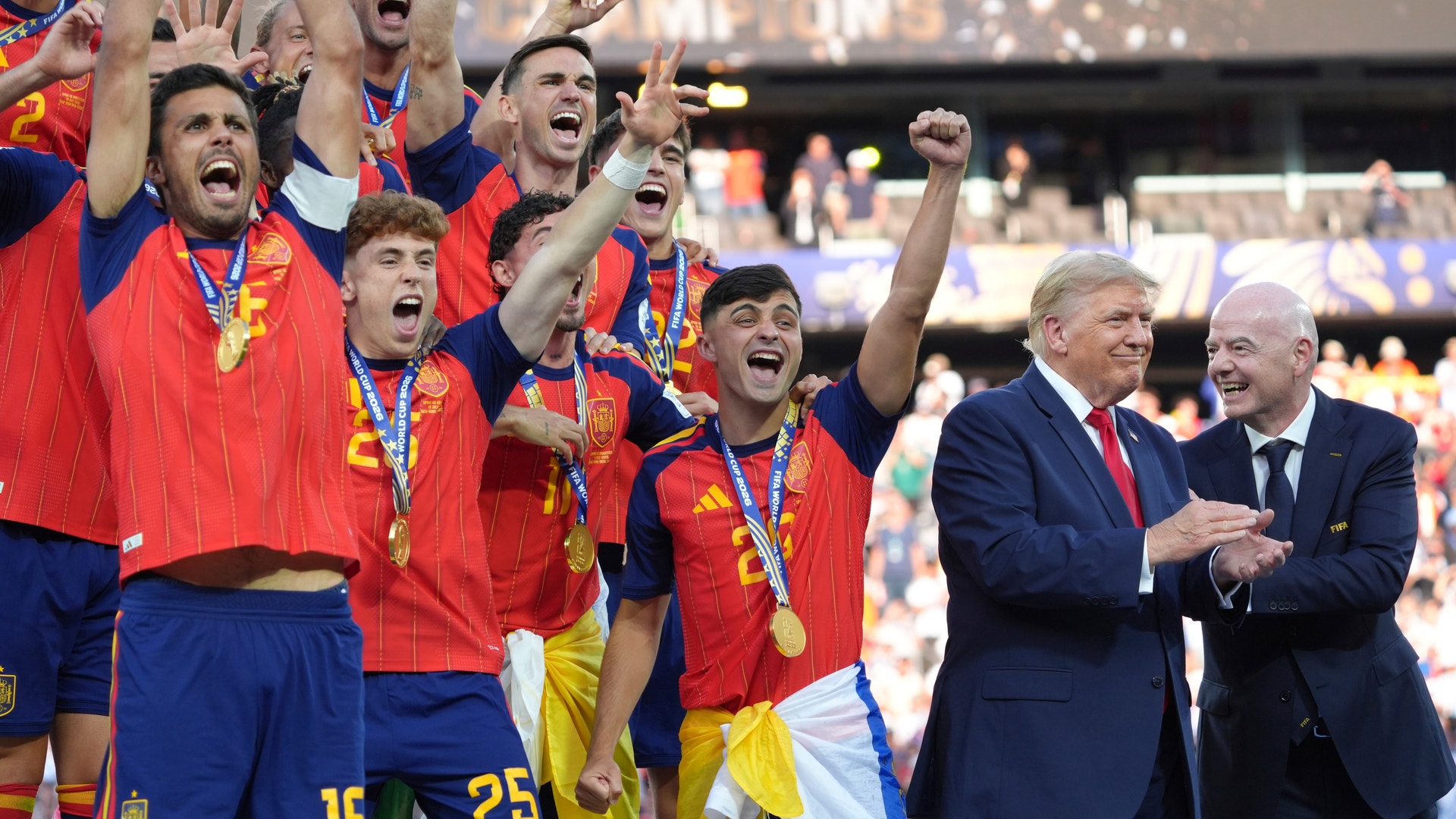 Fotbollsspelare i röda och blå landslagströjor med guldmedaljer jublar på en stadion efter en seger. Två män i kostymer står bredvid och ser glada ut.