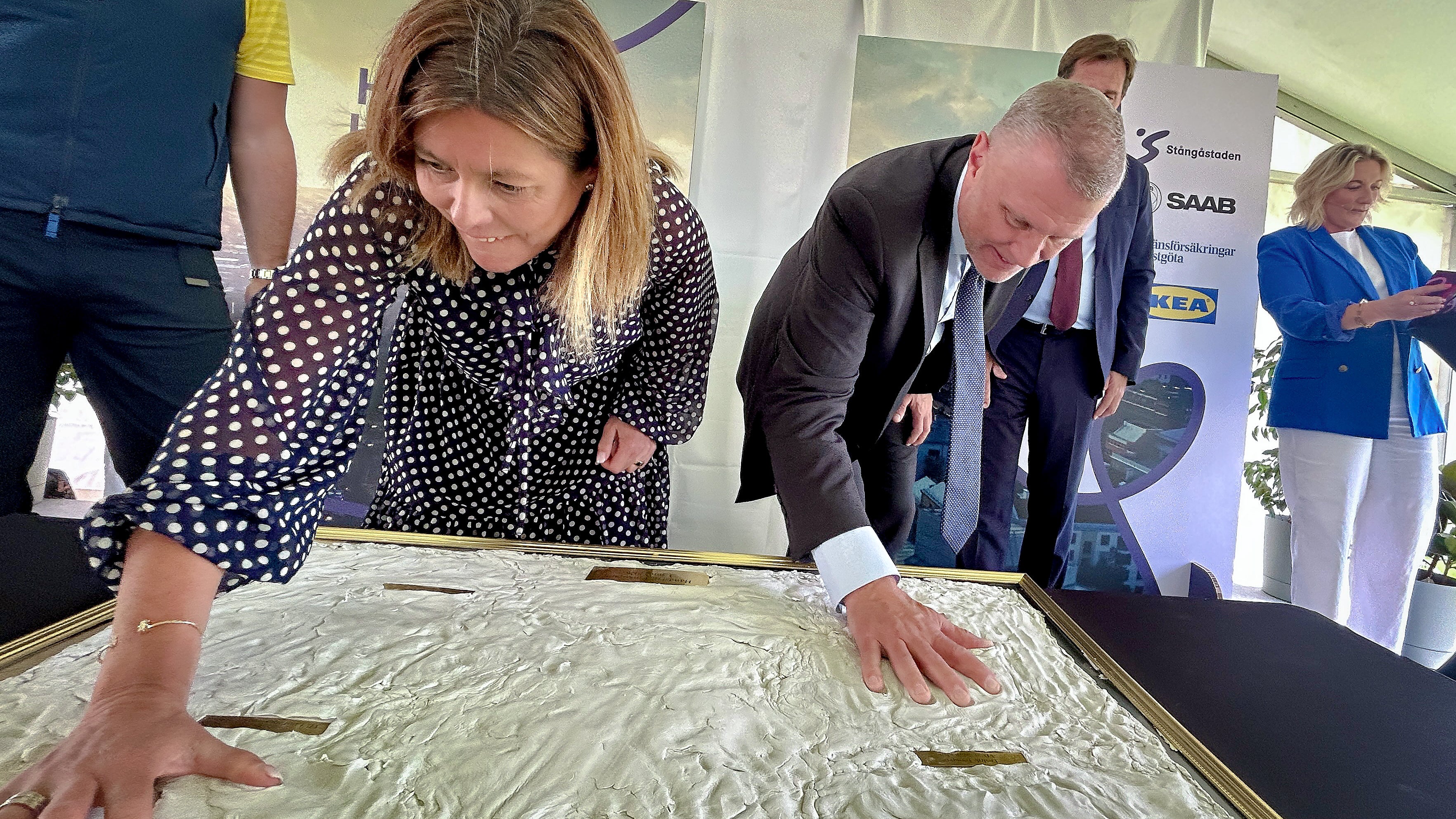 Flera personer i formell klädsel står omkring ett bord och undersöker en modell eller karta tillsammans vid ett evenemang.