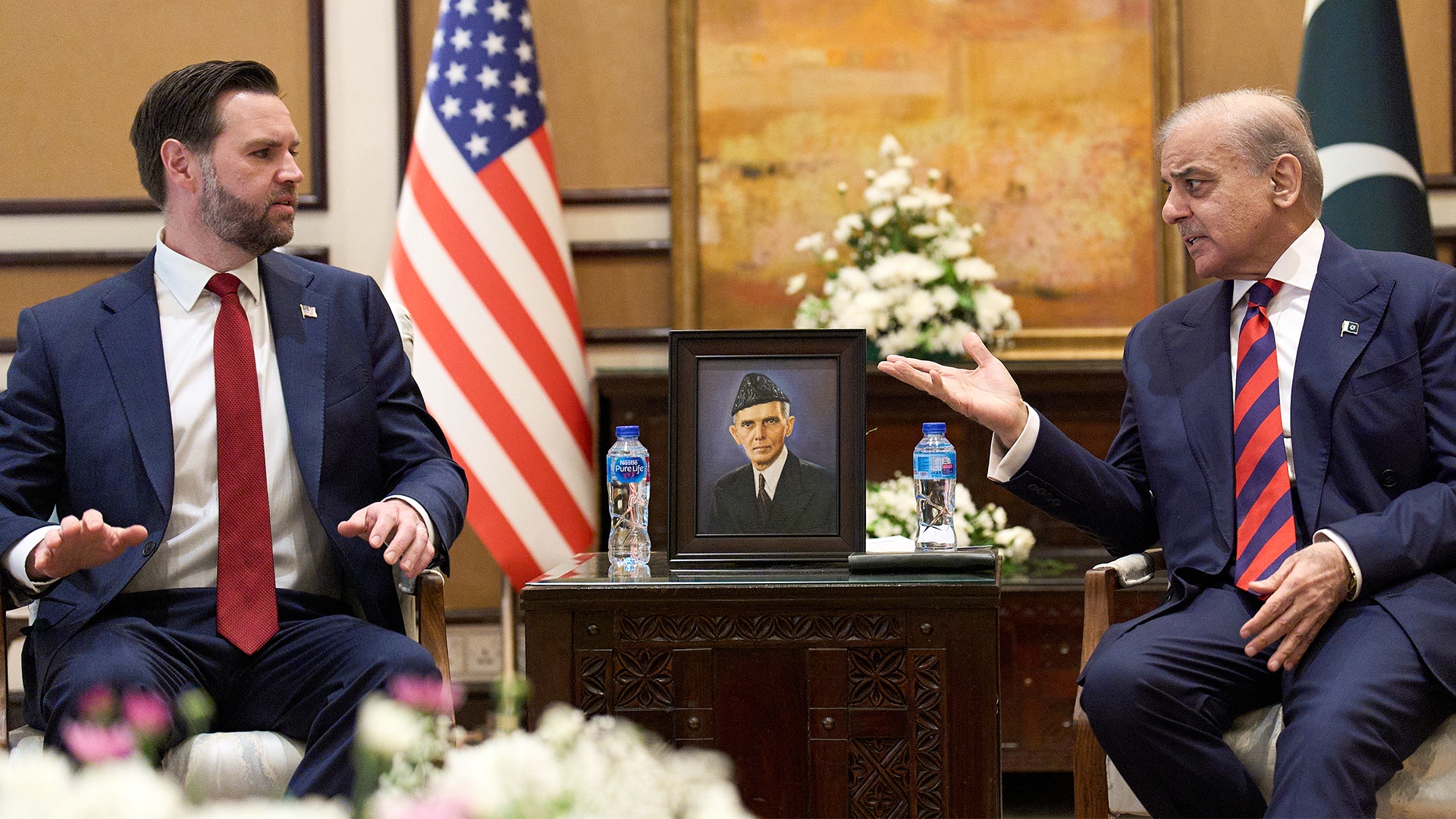 Två män i kostym sitter vid ett skrivbord under ett möte. En amerikansk flagga syns i bakgrunden tillsammans med en porträtt och blommor.