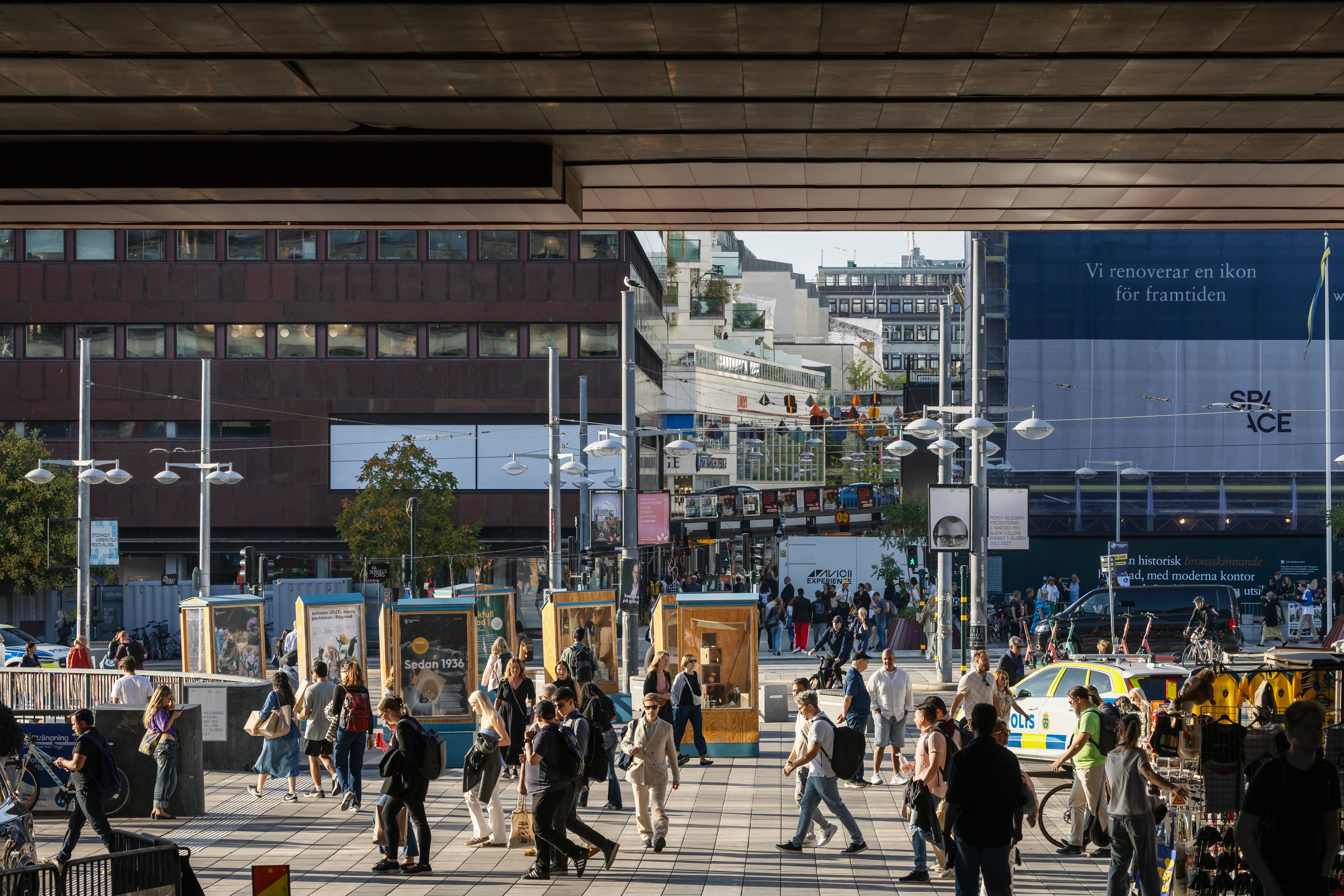 Många människor på ett öppet torg omgivet av moderna byggnader och skyltar.