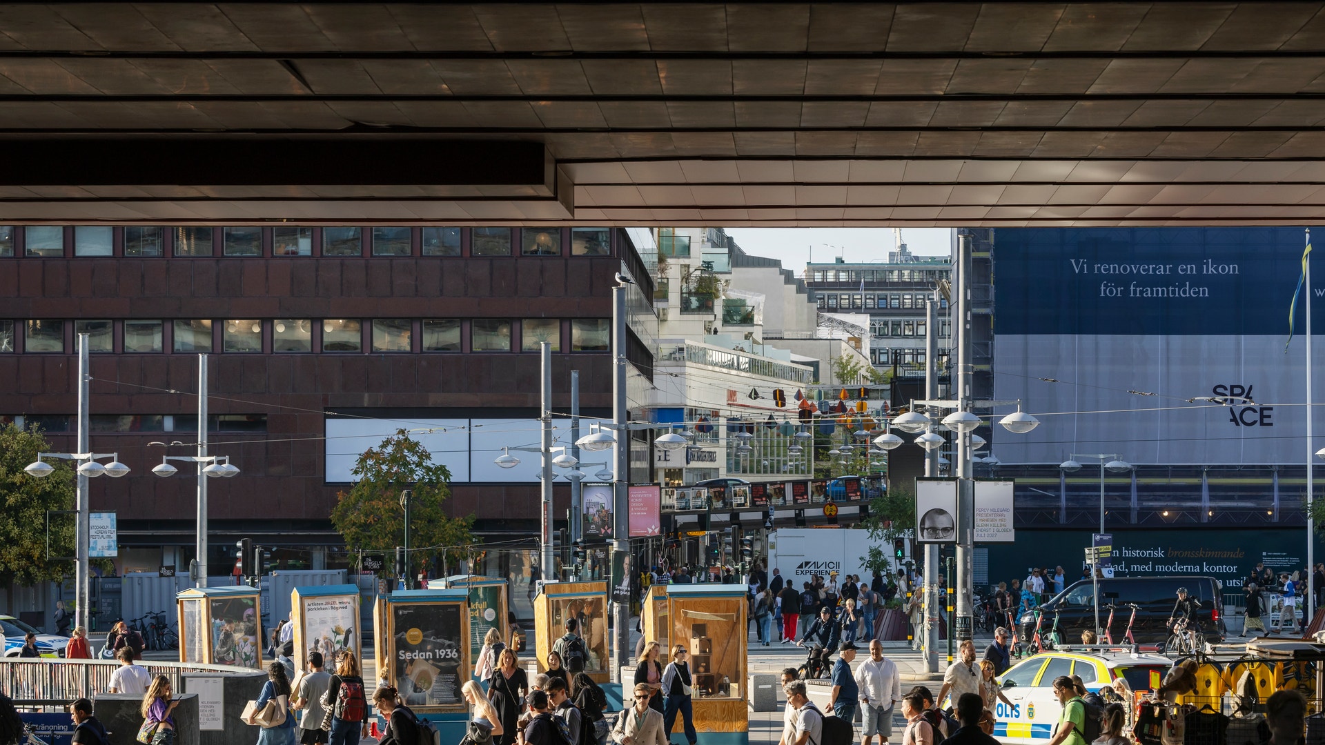 Många människor på ett öppet torg omgivet av moderna byggnader och skyltar.