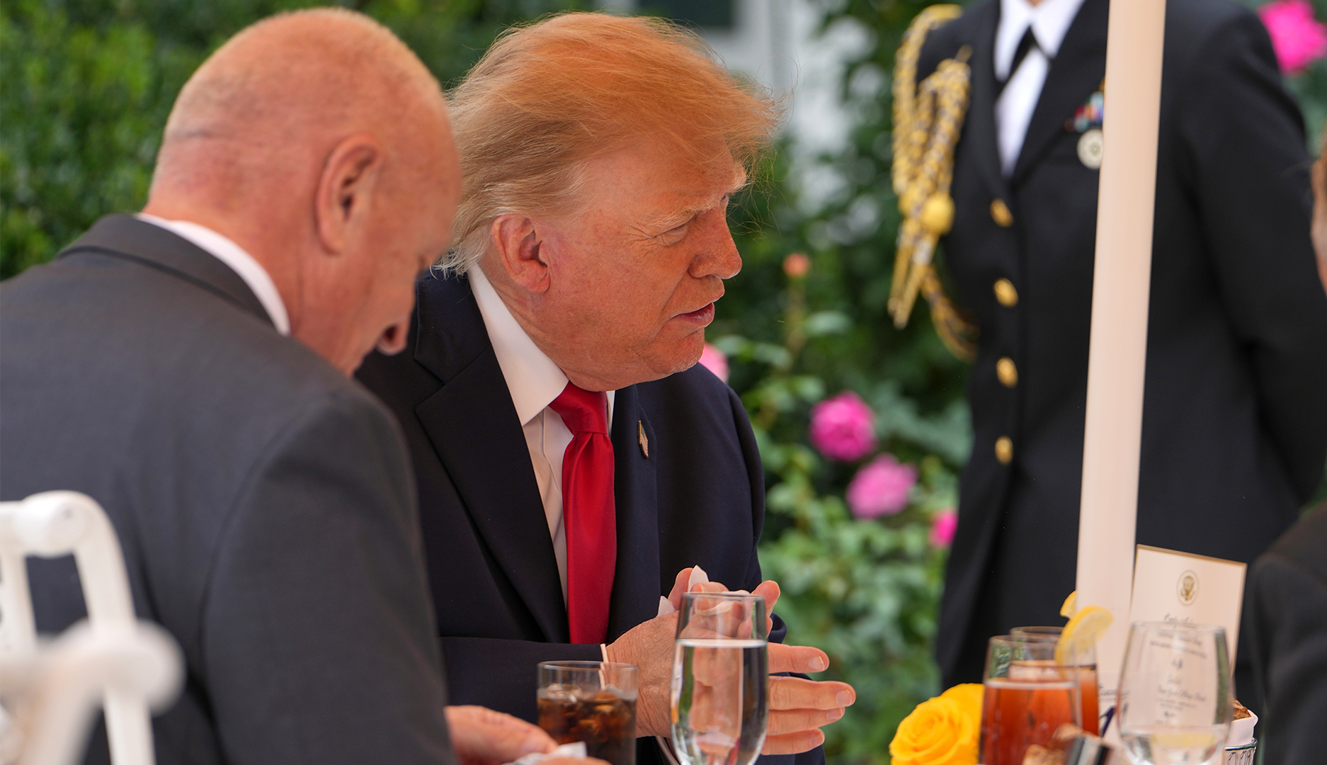 Donald Trump i mörk kostym och röd slips sitter vid ett middagsbord utomhus och samtalar med en man i förgrunden.