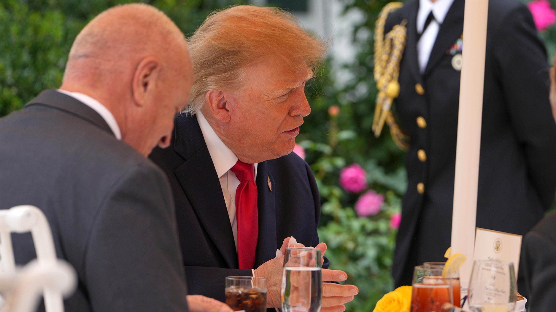Donald Trump i mörk kostym och röd slips sitter vid ett middagsbord utomhus och samtalar med en man i förgrunden.