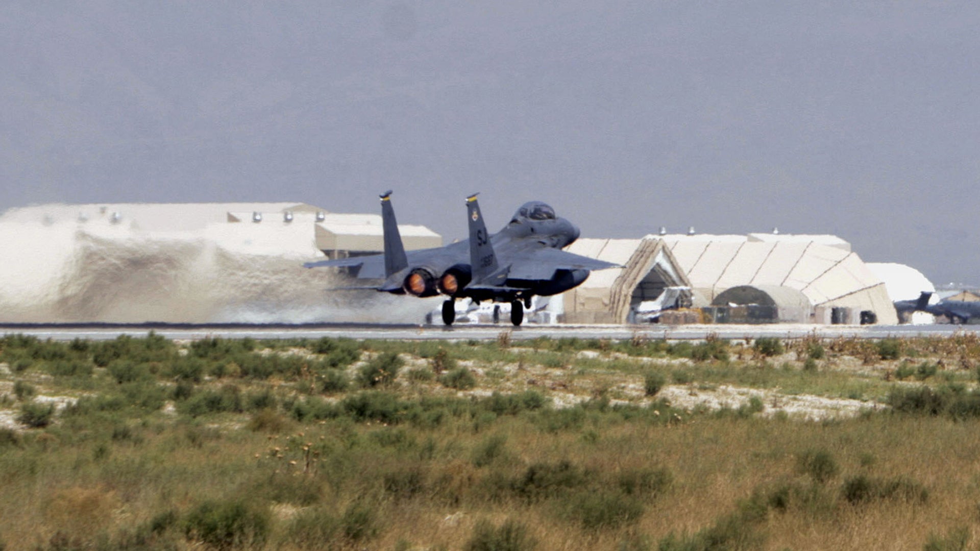 En militär jaktplan på en flygbas med hangarer i bakgrunden.