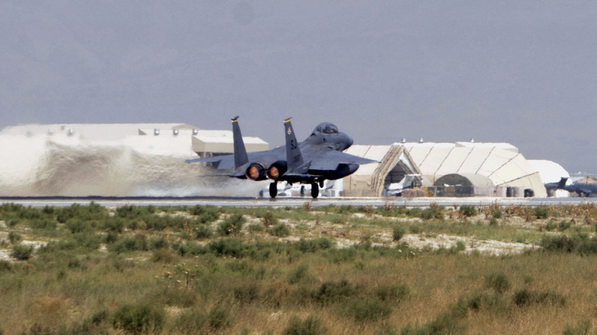 En militär jaktplan på en flygbas med hangarer i bakgrunden.