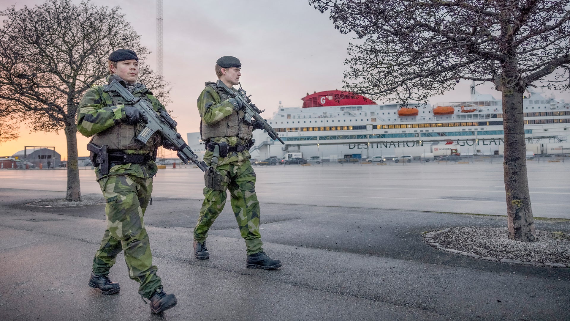 Två soldater i kamouflageuniformer patruller på en kaj vid en hamn. Ett stort passagerarfartyg ligger förtöjt i bakgrunden.