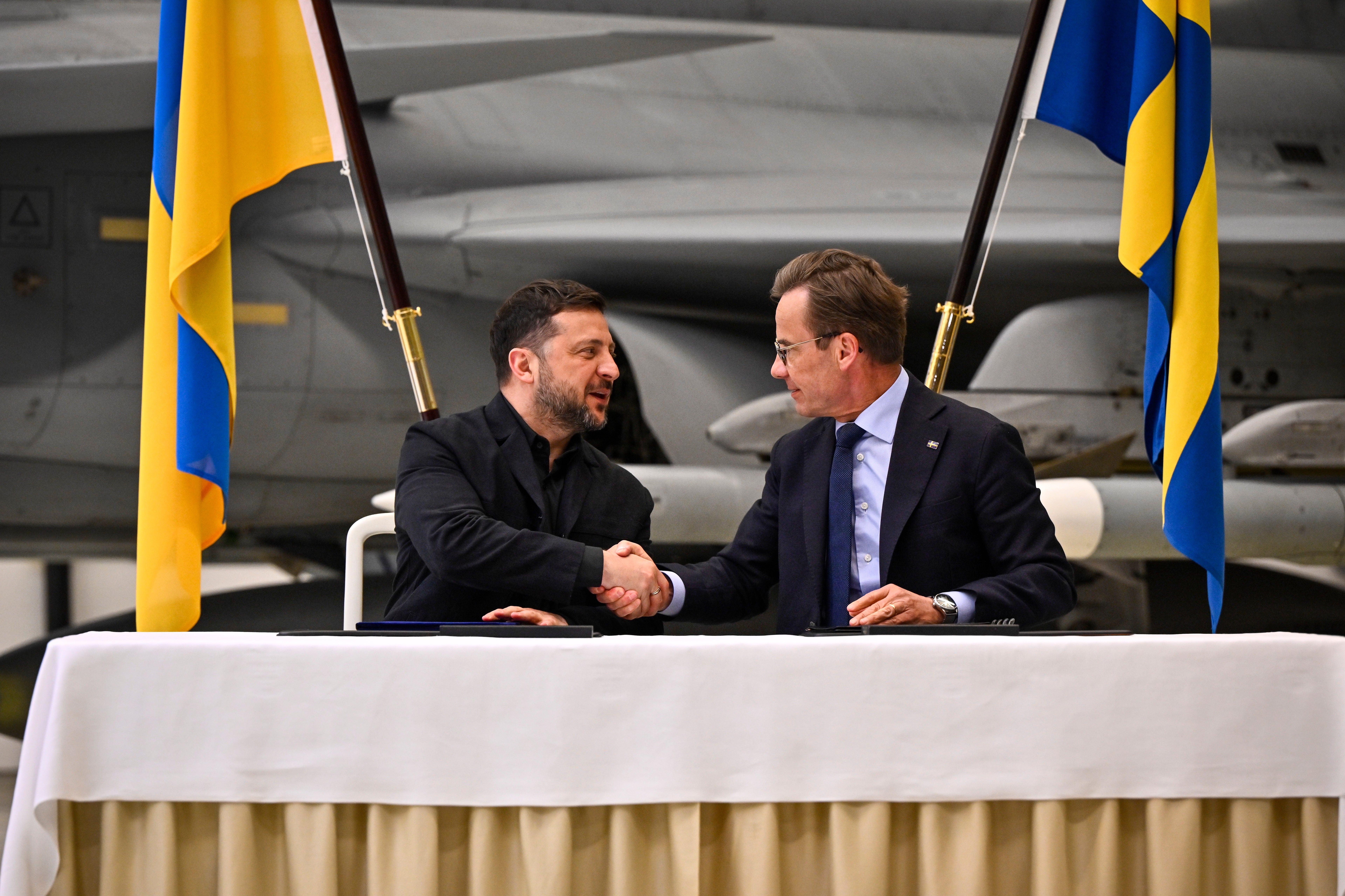 Två män skakar hand vid ett officiellt möte framför svenska flaggor.