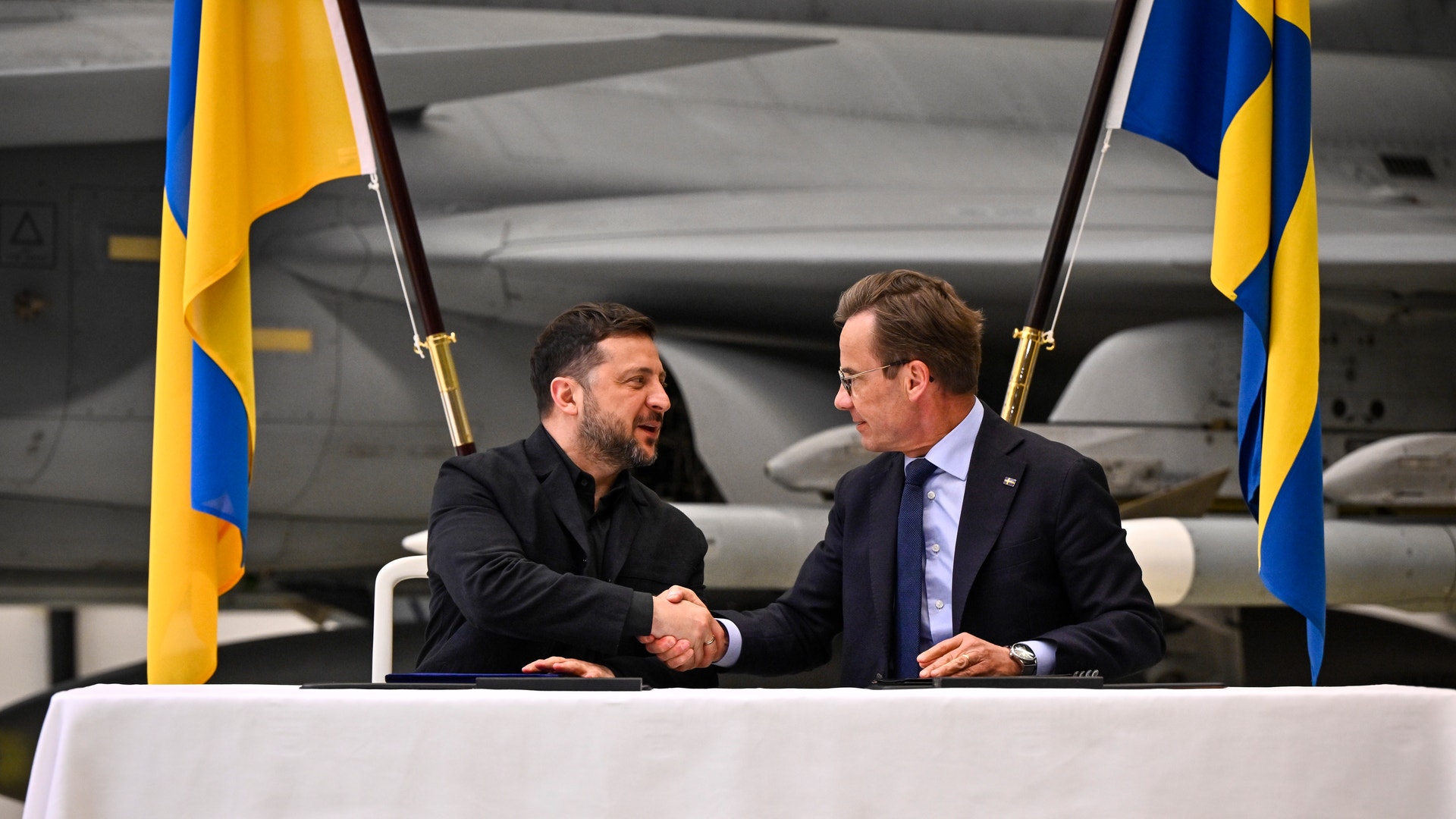 Två män skakar hand vid ett officiellt möte framför svenska flaggor.