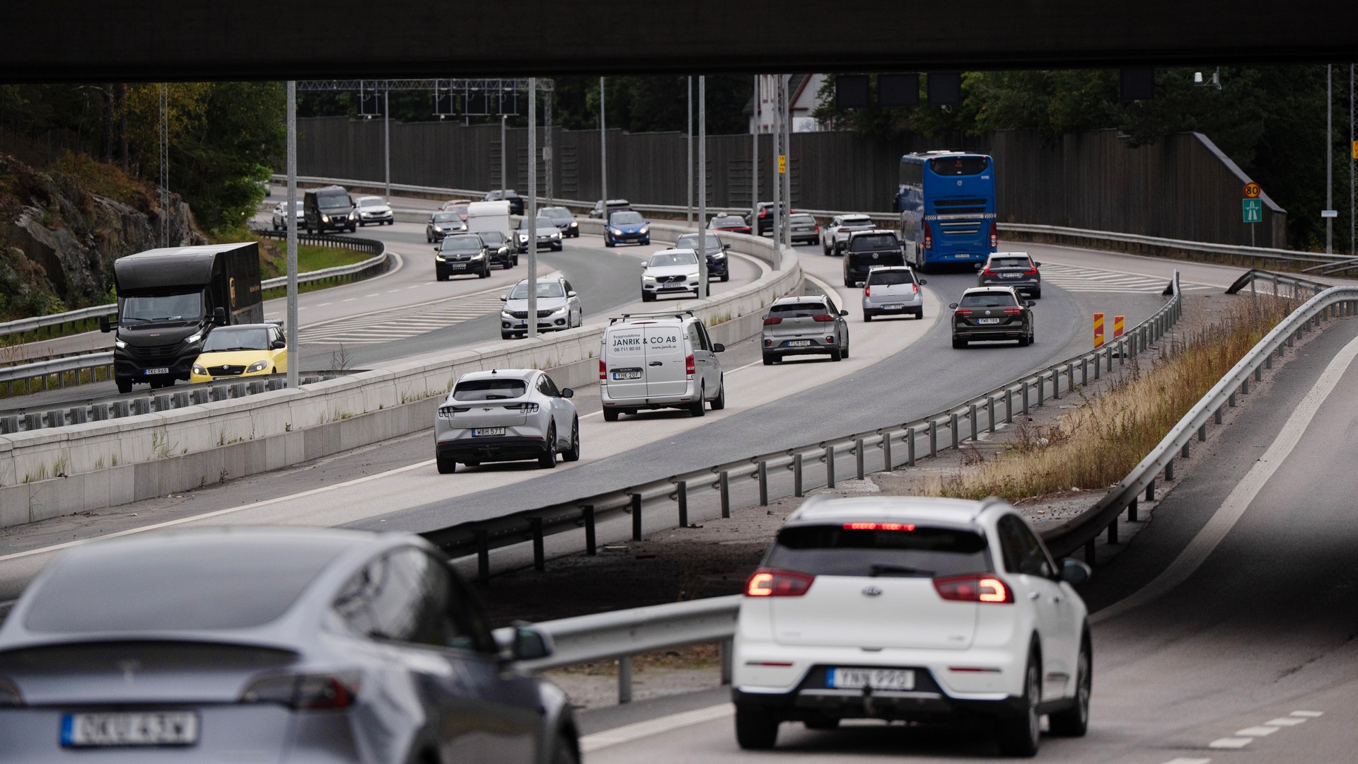 Trafikscen på en motorväg med flera körfält och olika fordon som bilar, lastbilar och bussar under en bro.
