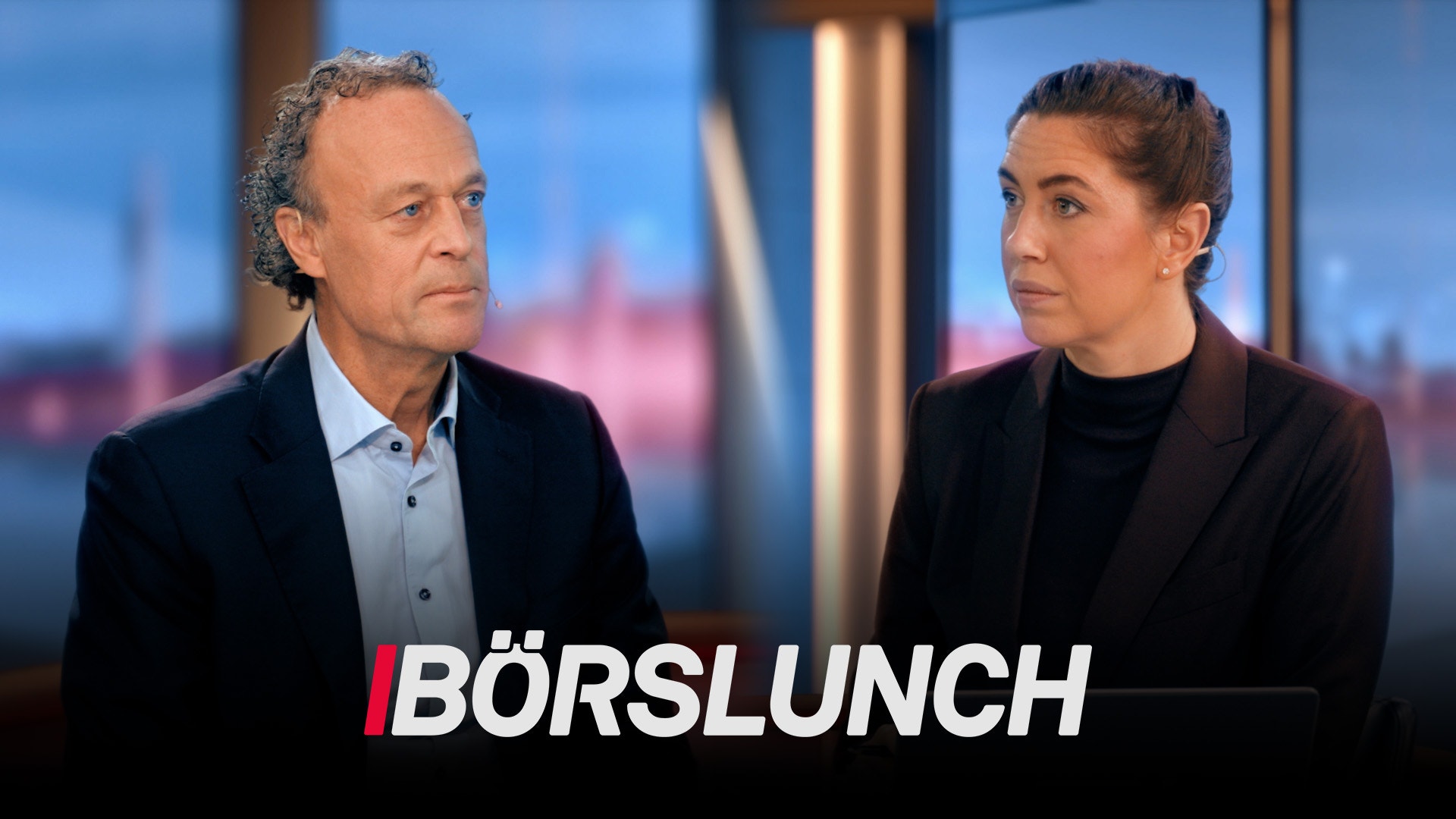 En man och en kvinna i affärskläder sitter mitt emot varandra i en studiomiljö med en stadshorisont i bakgrunden. Mannen bär en mörk blazer och ljusblå skjorta, kvinnan bär en svart blazer. De verkar diskutera något allvarligt. Texten "BÖRSLUNCH" visas längst ned på bilden.