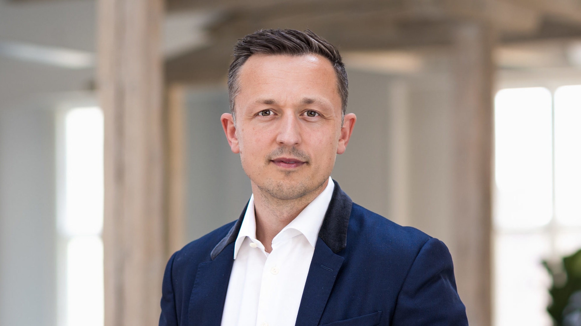 Porträtt av en man i mörkblå kavaj och vit skjorta i en modern kontorsmiljö.