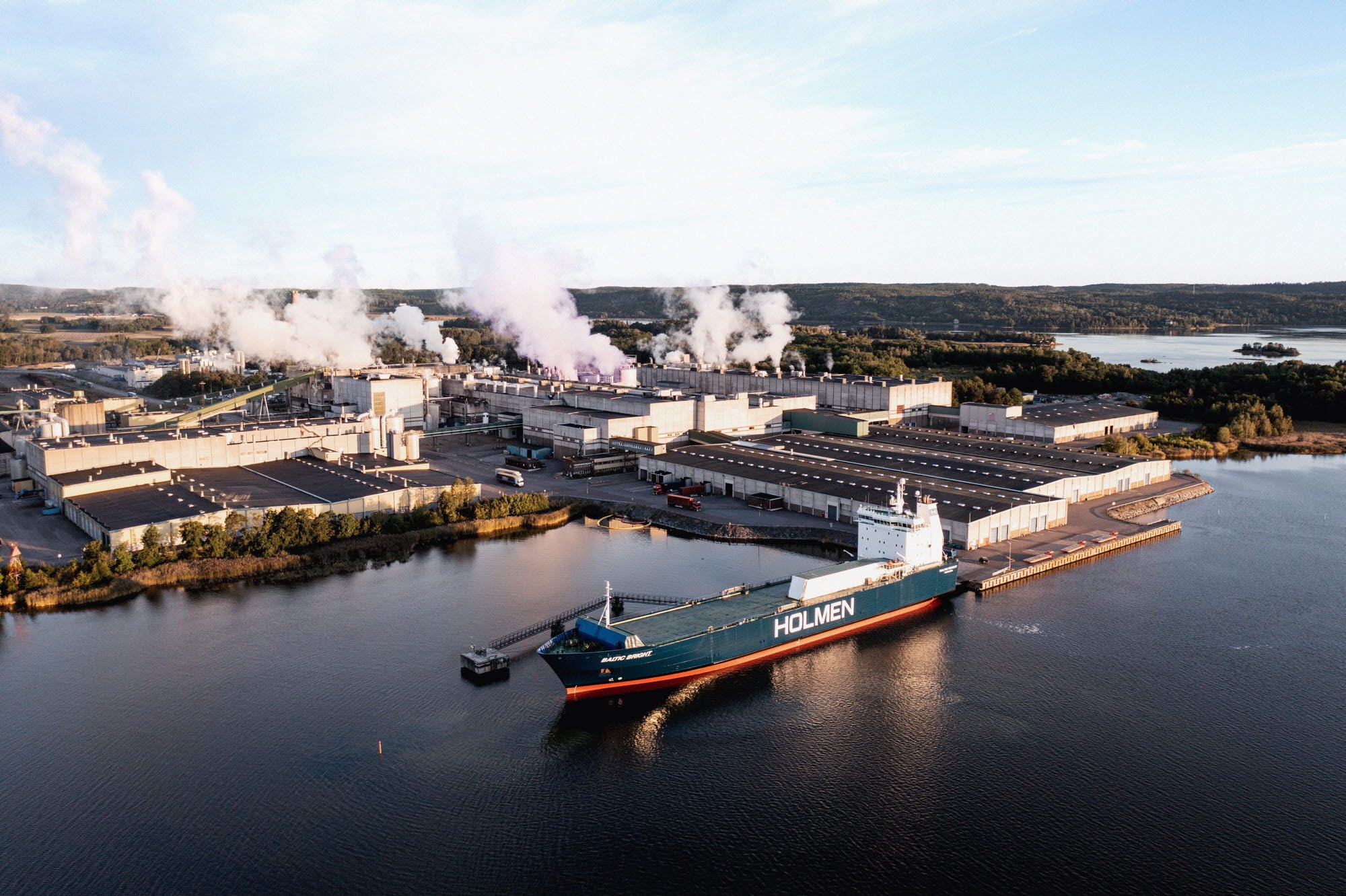 Flygfoto av Holmen Braviken-fabriken vid vattnet. Ett stort industrifartyg ligger dockat vid fabrikens kaj. Fabrikens skorstenar släpper ut rök. Området är omgivet av skog och vatten.