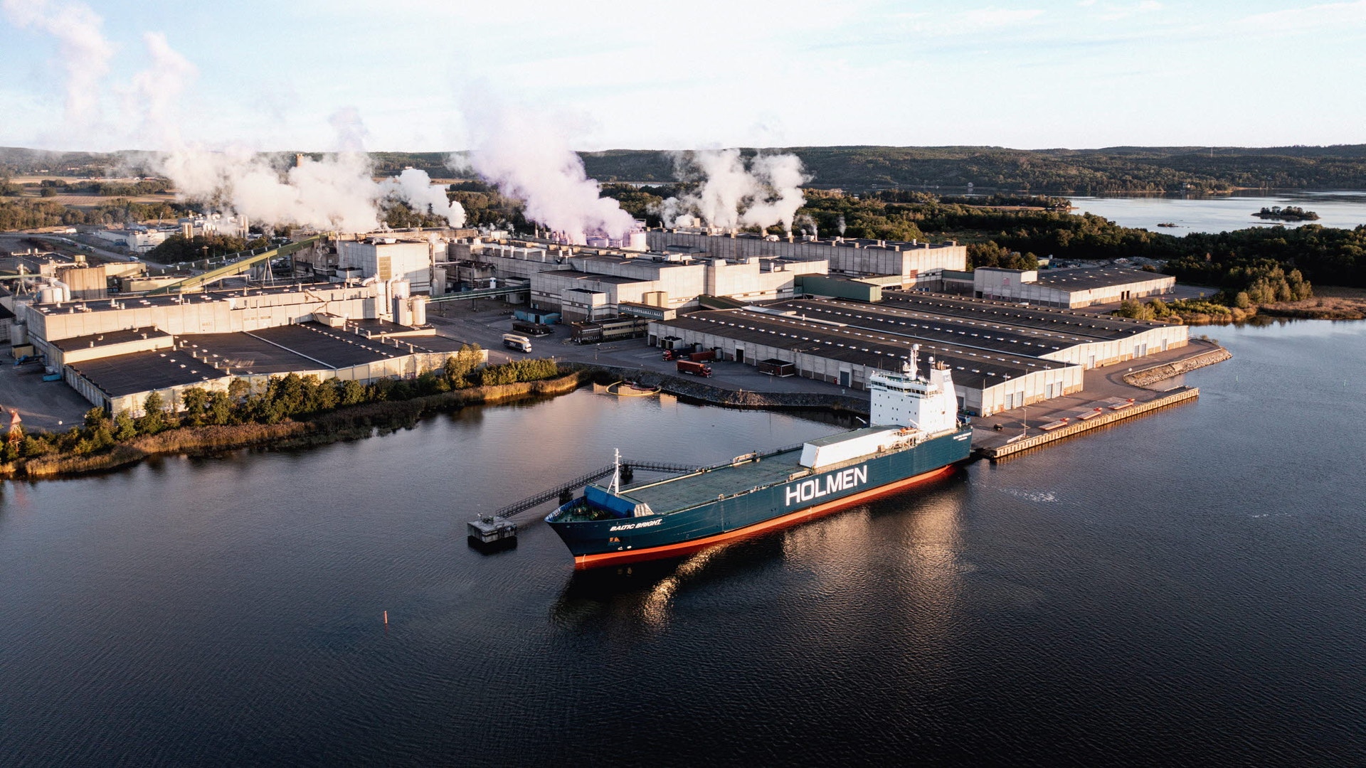 Flygfoto av Holmen Braviken-fabriken vid vattnet. Ett stort industrifartyg ligger dockat vid fabrikens kaj. Fabrikens skorstenar släpper ut rök. Området är omgivet av skog och vatten.