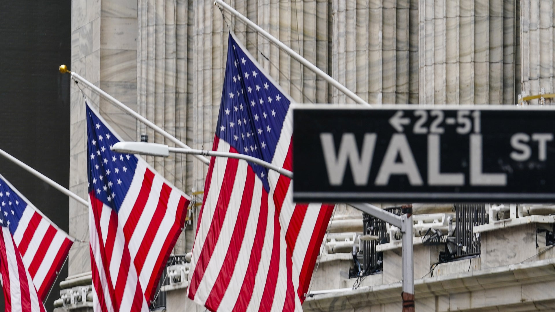 Amerikanska flaggor hängande framför en byggnad på Wall Street i New York, med en Wall Street-skylt synlig i bakgrunden.