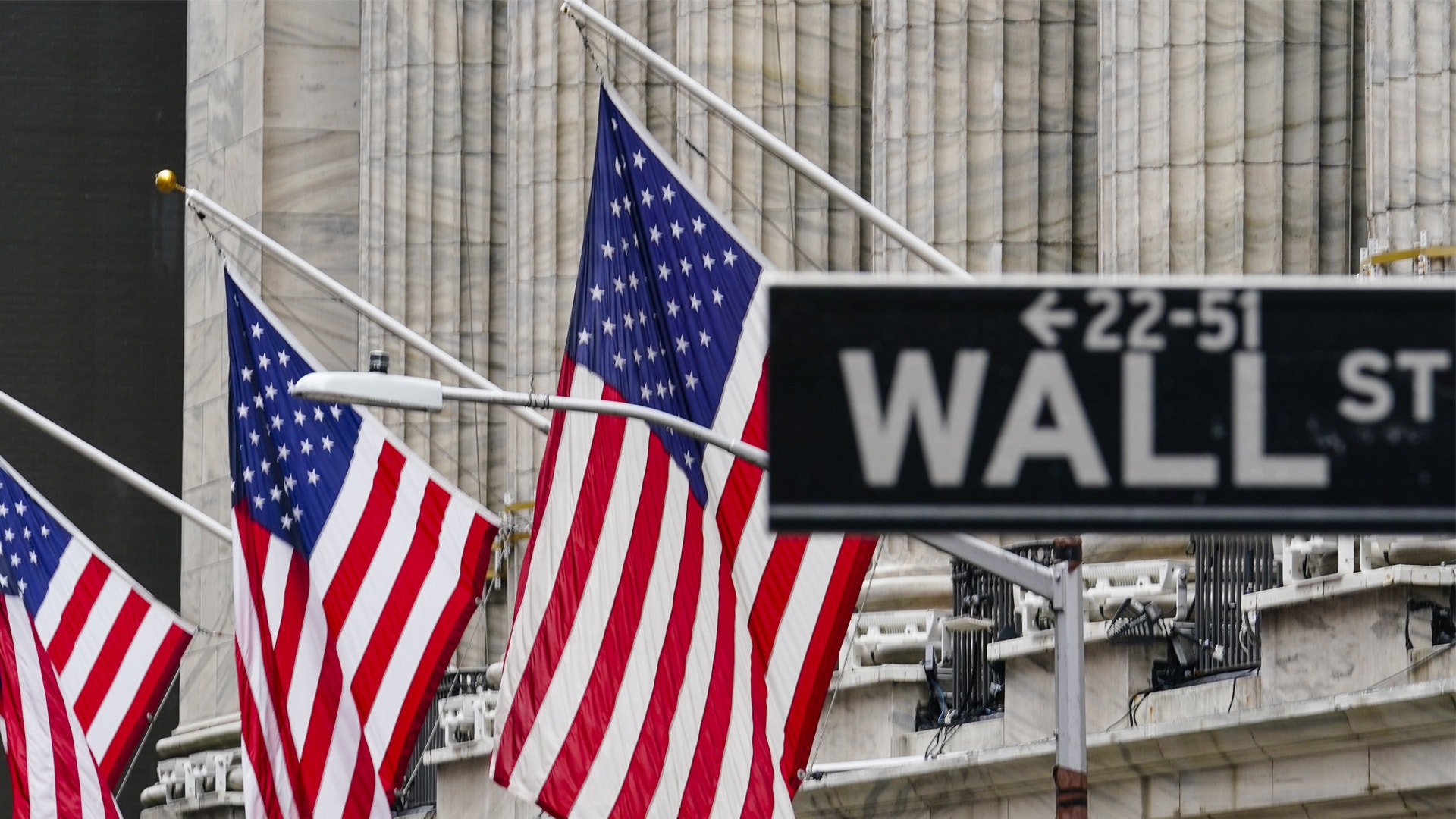 Amerikanska flaggor hängande framför en byggnad på Wall Street i New York, med en Wall Street-skylt synlig i bakgrunden.
