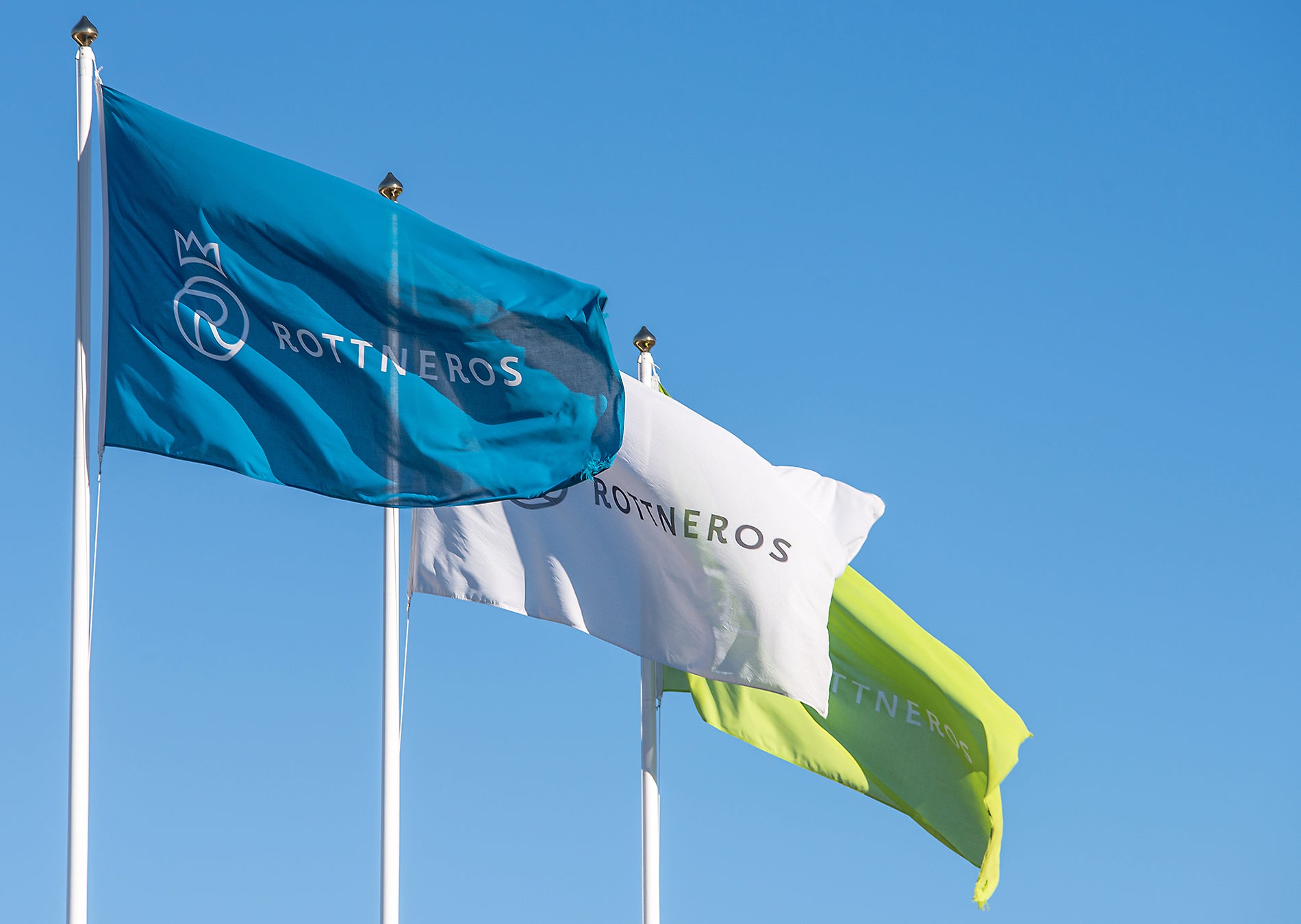 Tre flaggor som vajar mot en blå himmel. Flaggorna visar Rottneros logotyp och märke.